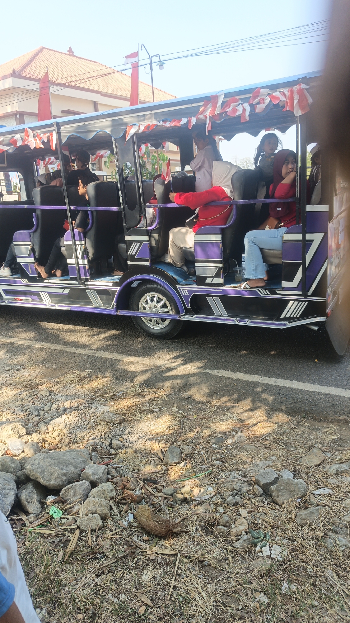 In Kandangmas, Indonesia, a group of people are riding in a open-air vehicle decorated with red and white Indonesian flags. The vehicle appears to be a modified jeep or a similar type of transport. Several individuals, including women and children, are seated inside. The vehicle has a distinctive purple and silver design along its side. In the background, a building with a red-tiled roof and flagpoles is visible. The scene takes place on a road with a rocky and dry grassy shoulder. The lighting suggests it is daytime, with sunlight casting shadows.