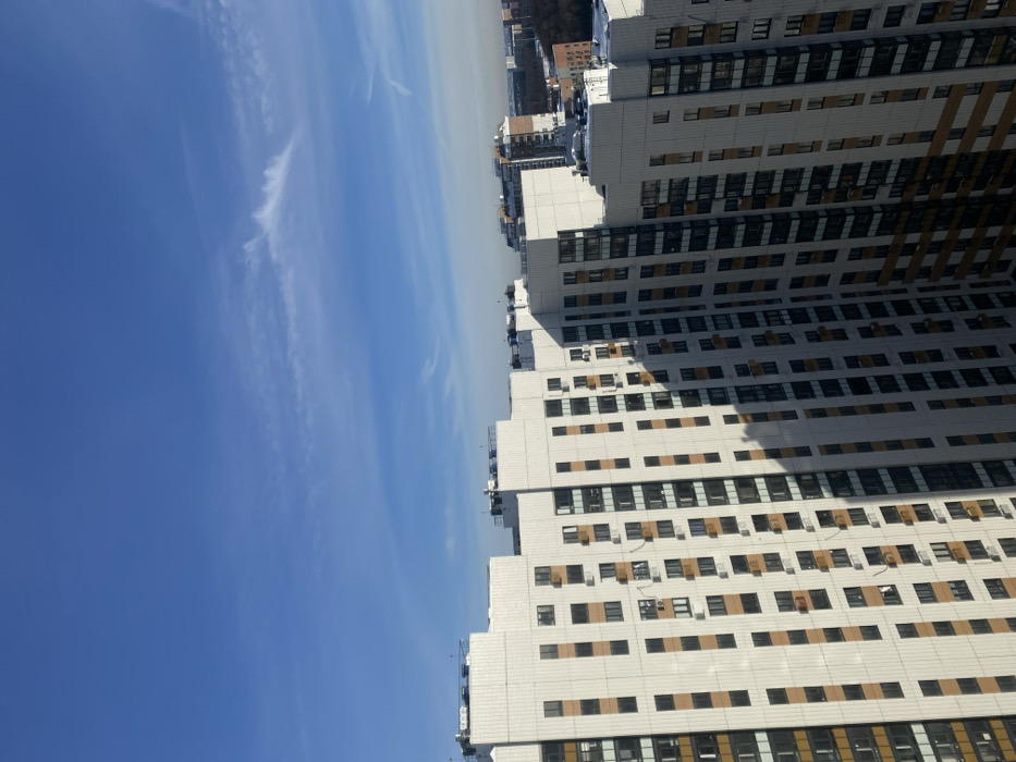 The image presents an urban landscape in Bittsa, Russia, captured from an elevated perspective. The upper portion of the frame is occupied by a clear, bright blue sky, featuring scattered, wispy white cirrus clouds, with a notable elongated cloud formation stretching across the upper right. The lower half displays a cluster of modern, high-rise residential buildings. These structures possess light-colored, possibly off-white, facades interspersed with dark-framed windows arranged in a grid pattern and vertical sections of light brown paneling. Numerous external air conditioning units are mounted on various building walls. Strong sunlight casts defined shadows from one building onto another, particularly on the right side of the image, obscuring parts of a facade in shade. Building rooftops are flat and feature visible mechanical equipment. In the far distance, additional city infrastructure is visible, alongside a segment of what appears to be a forested area. Some distant rooftops exhibit traces of snow. No human figures, discernible actions, or interactions are present in the scene.