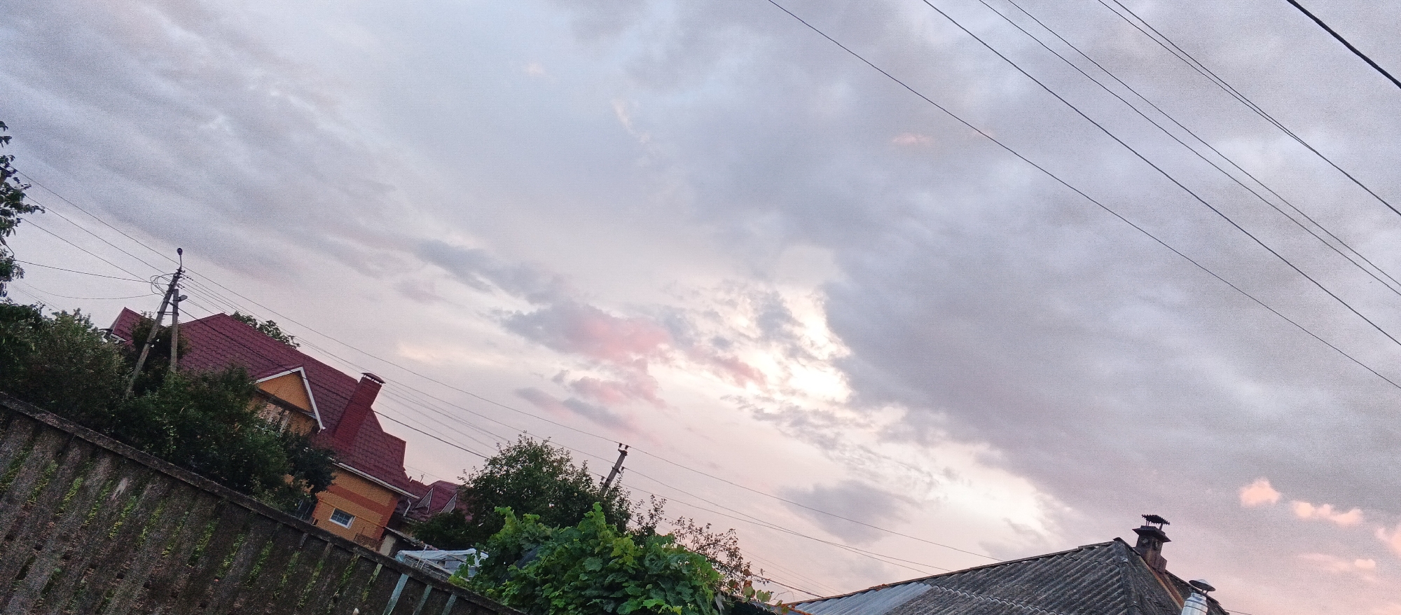 The image depicts a suburban scene in Smila, Ukraine, characterized by a cloudy sky at dusk. In the foreground, a weathered, grey fence runs diagonally across the frame, partially obscured by lush green foliage. Behind the fence, a two-story house with a distinctive red tiled roof and yellow siding is visible, surrounded by trees. Electrical wires crisscross the sky, supported by utility poles. To the right, another roofline with a chimney is visible, suggesting more residential structures. The sky is filled with a mix of grey and lighter clouds, with subtle hints of pink and orange light indicating the setting sun. The overall impression is one of a quiet, residential neighborhood as day transitions into night.