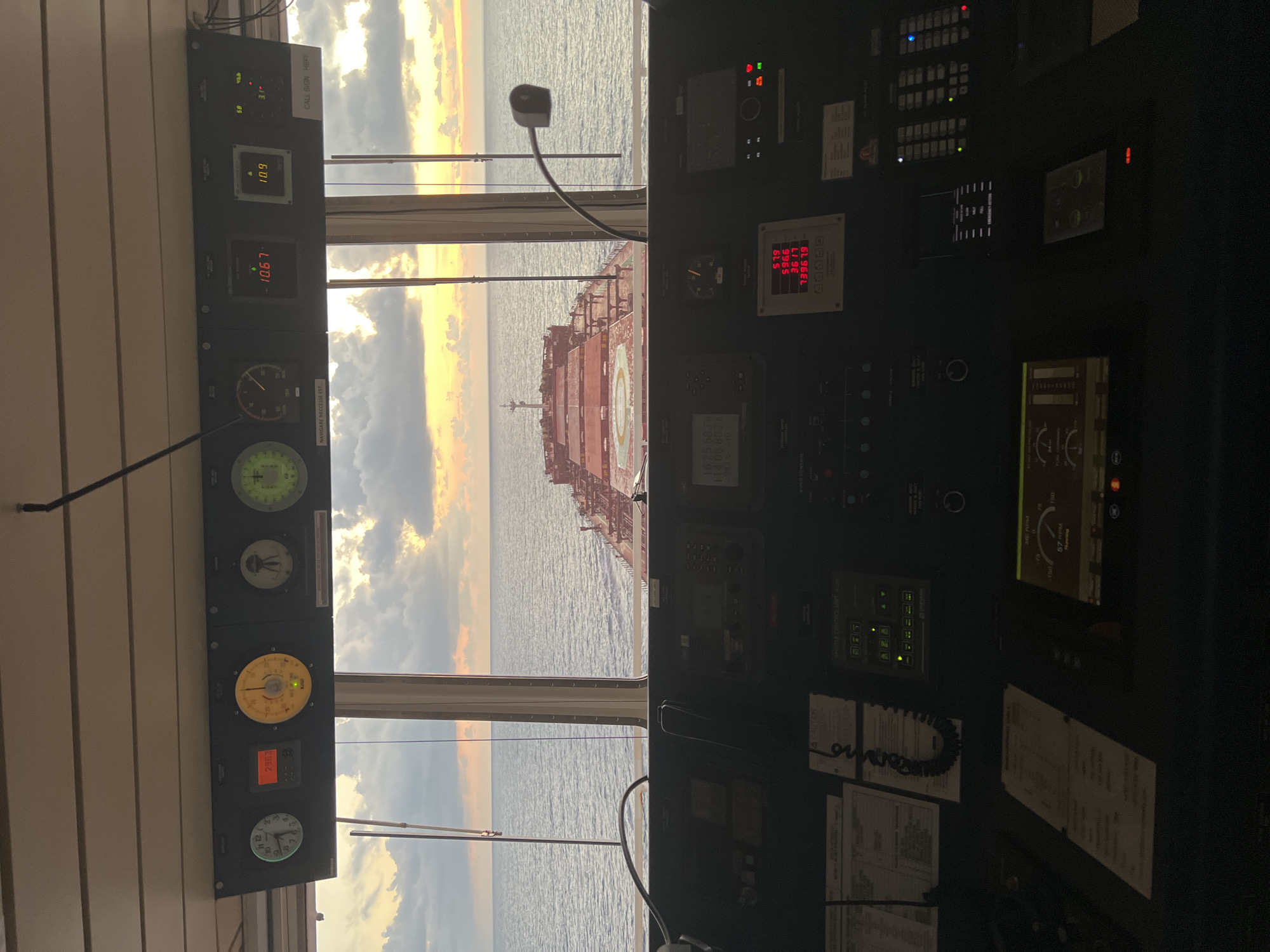 The image shows the interior of a ship's bridge, looking out at the ocean at sunset. The bridge is filled with various control panels, gauges, and screens. In the foreground, there are multiple consoles with buttons, switches, and displays. A steering wheel is visible, as well as a large window overlooking the sea. The window reveals a cargo ship sailing away in the distance, silhouetted against a sky filled with orange and yellow clouds. The overall scene depicts a functional and busy maritime environment.