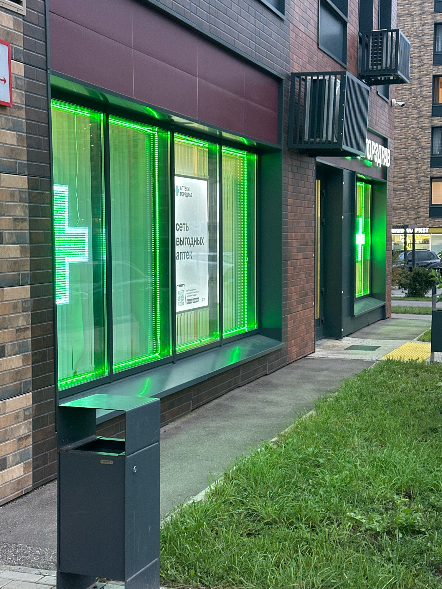 The image depicts the exterior of a pharmacy in Vidnoye, Russia. The pharmacy features large windows illuminated with bright green neon lights, forming a cross shape on the left window and vertical strips on the right. A sign with Russian text and a QR code is visible through the window on the right. To the left of the pharmacy entrance, there is a dark gray outdoor trash receptacle. The pharmacy is situated on a street with a sidewalk and a strip of green grass. The building's facade is made of dark brick and a maroon paneling. In the background, another building is visible, along with trees and a parked car. The lighting suggests it is either dusk or an overcast day.