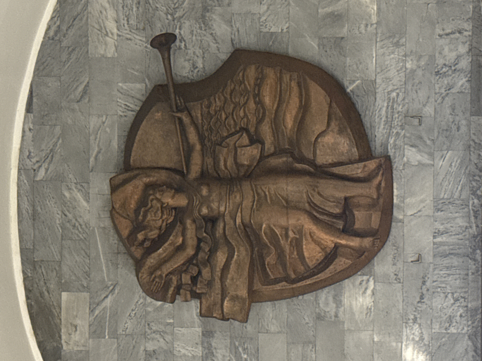 A low-angle, medium shot captures a bronze-colored relief sculpture mounted on a gray, stone-tiled wall. The sculpture features a recumbent, draped figure, possibly female, with flowing hair and a hand resting on a stack of what appear to be bricks. A long, horn-like instrument rises from behind the figure. The sculpture is mounted on a semi-circular or shield-shaped backing. The setting appears to be an interior space with a minimalist aesthetic, possibly a public building. The sculpture is located at the Finland Railway Station in Saint Petersburg, Russia, symbolizing the arrival of Lenin.