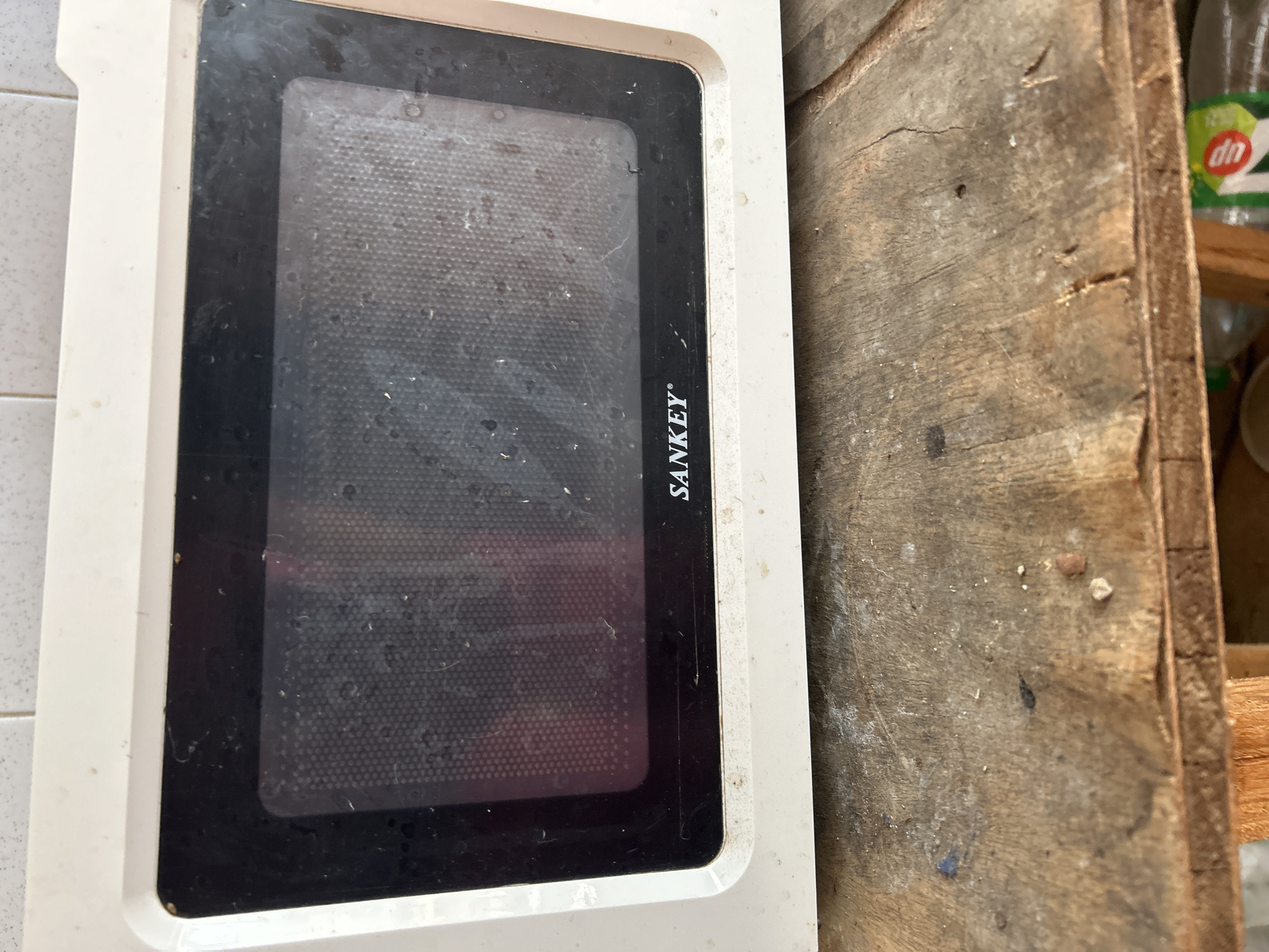 The image shows a close-up of a dirty white Sankey microwave oven sitting on a weathered wooden surface. The microwave door is black with a visible grid pattern inside, and there are water spots and smudges on the glass. The Sankey logo is visible below the glass. The wooden surface appears to be part of a table or shelf, showing cracks and debris. The background is out of focus, but suggests a tiled wall behind the microwave. There is no indication of the city of Santa Ana, Venezuela in the image.