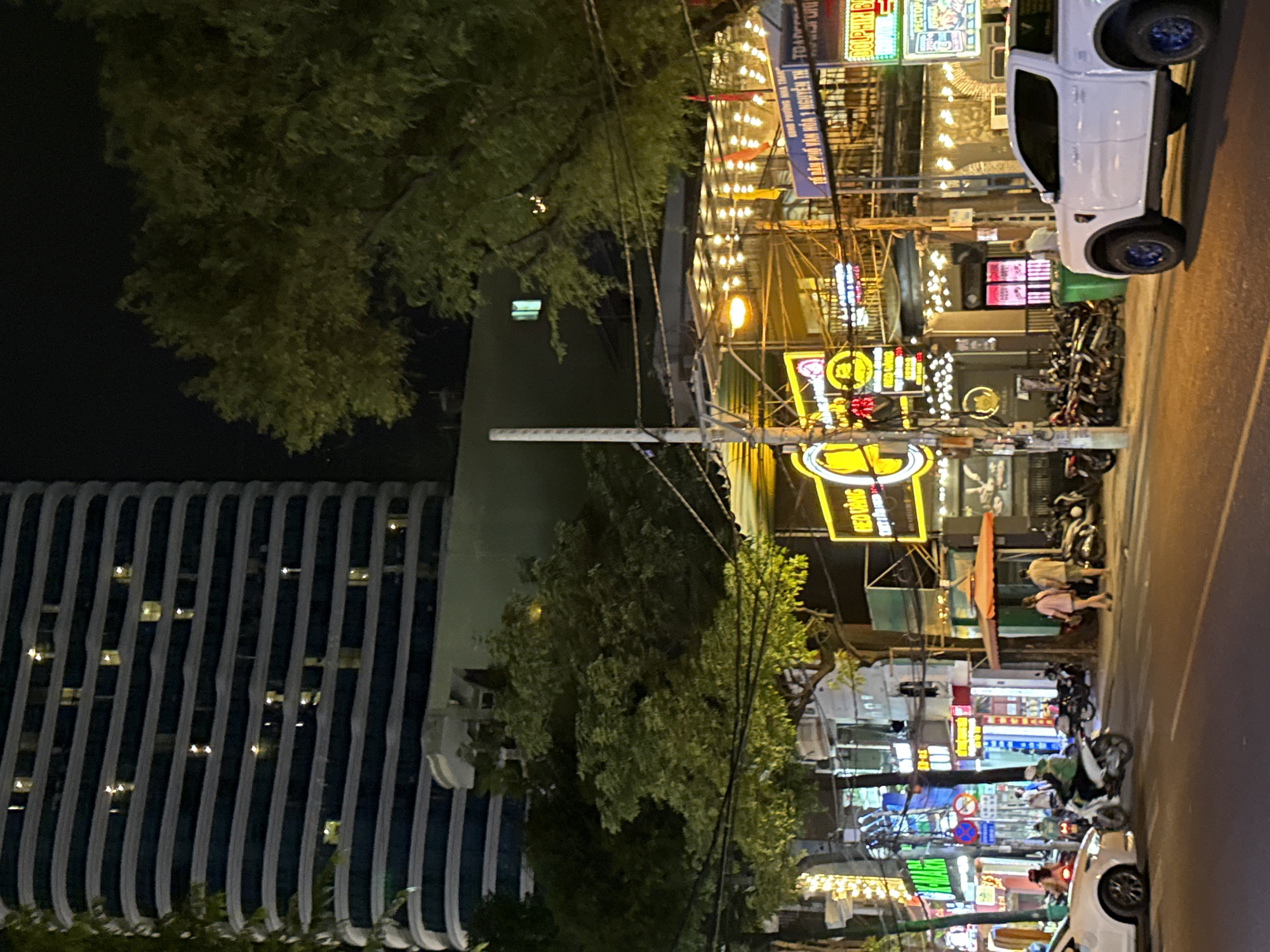 This image depicts a nighttime street scene in Nha Trang, Vietnam, focusing on a busy commercial area. A tall, modern building with horizontal lines and illuminated windows dominates the background. In the midground, a tree with dense foliage is illuminated by the vibrant neon signs of various businesses. These signs, written in Vietnamese, advertise restaurants and shops. The street is bustling with activity; motorcycles are parked along the sidewalk and a few are in motion, with riders visible. Pedestrians, including a couple walking across the street and a person standing near a green trash bin, are present. A white pickup truck with blue rims is parked on the right side of the road. The scene is characterized by the bright lights of the signs and building, creating a lively and somewhat chaotic urban atmosphere.