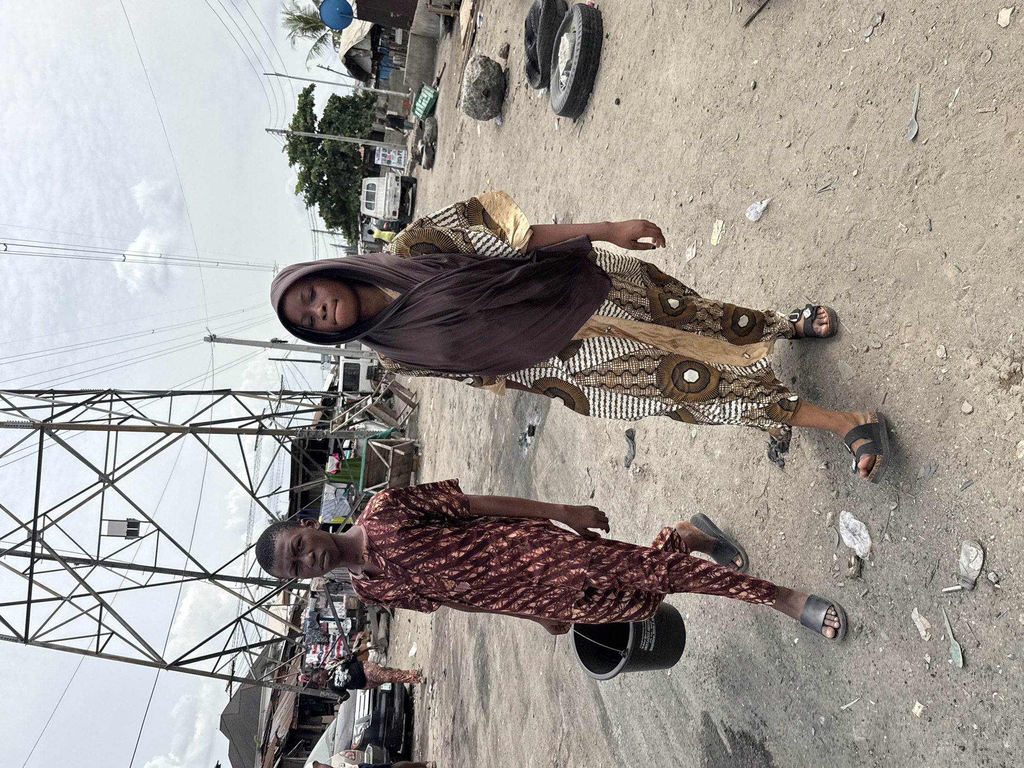 Two individuals are standing on a dirt road in Ado, Nigeria. The first individual is a woman wearing a tan and white patterned dress with a dark brown head covering. She is standing next to an electrical tower. The second individual is wearing a brown patterned dress and carrying a black bucket. The environment includes dirt roads, litter, and a view of buildings and vegetation in the background.