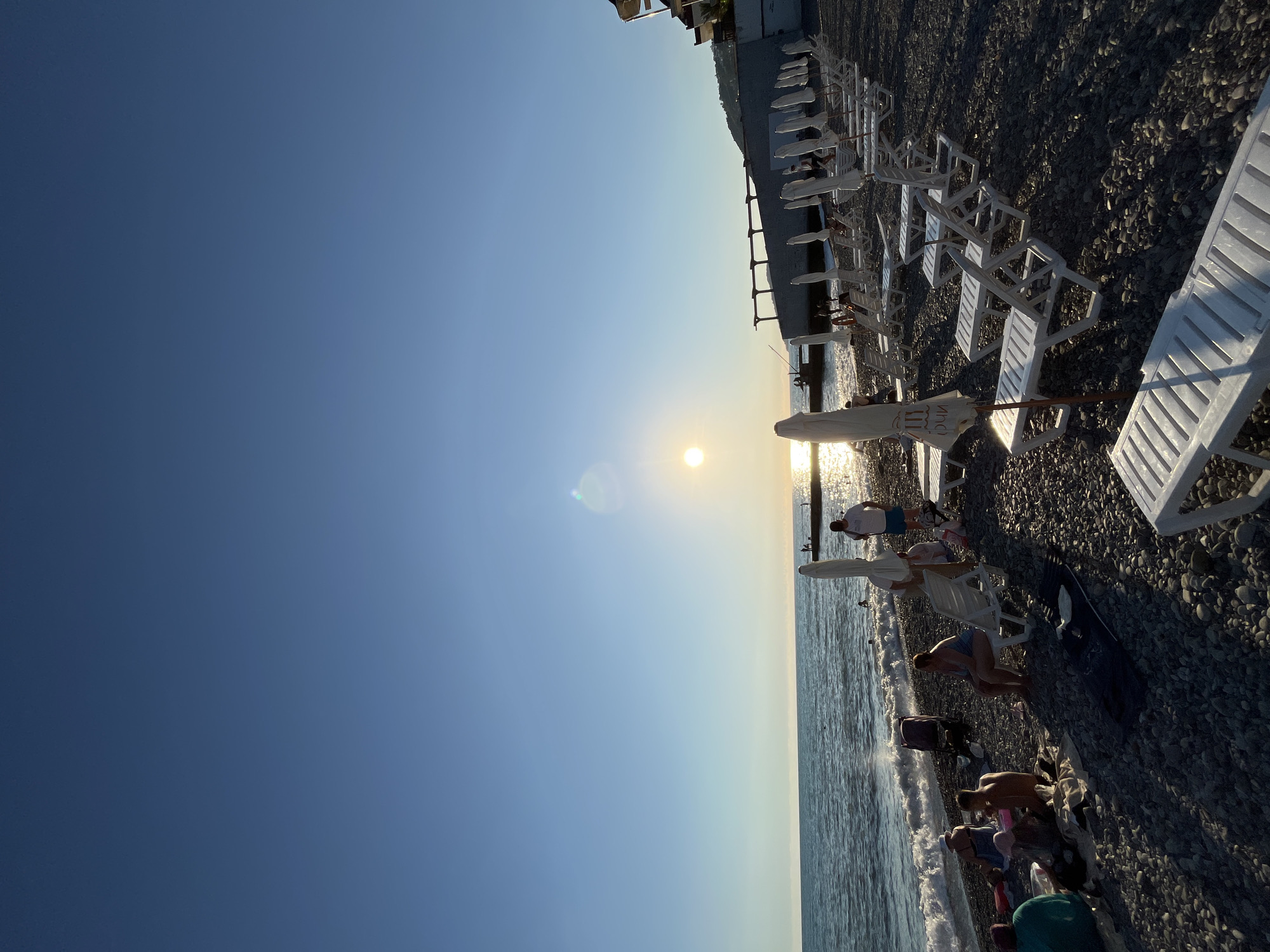 The photograph depicts a pebble beach in Sochi, Russia, during a sunset. The sun is low in the sky, casting a warm glow and creating a lens flare. The sea is calm with gentle waves lapping the shore. Rows of empty white lounge chairs and folded umbrellas are neatly arranged on the beach, suggesting a resort area. Several people are visible on the beach, some sitting on towels and loungers, while others are standing or walking near the water's edge. In the distance, a pier or breakwater extends into the sea, with a few figures on it. The background features a concrete structure with pergolas and a hillside with buildings, characteristic of the Sochi coastline. The sky is clear and blue, transitioning to a lighter hue near the horizon where the sun is setting. The overall atmosphere is serene and peaceful.