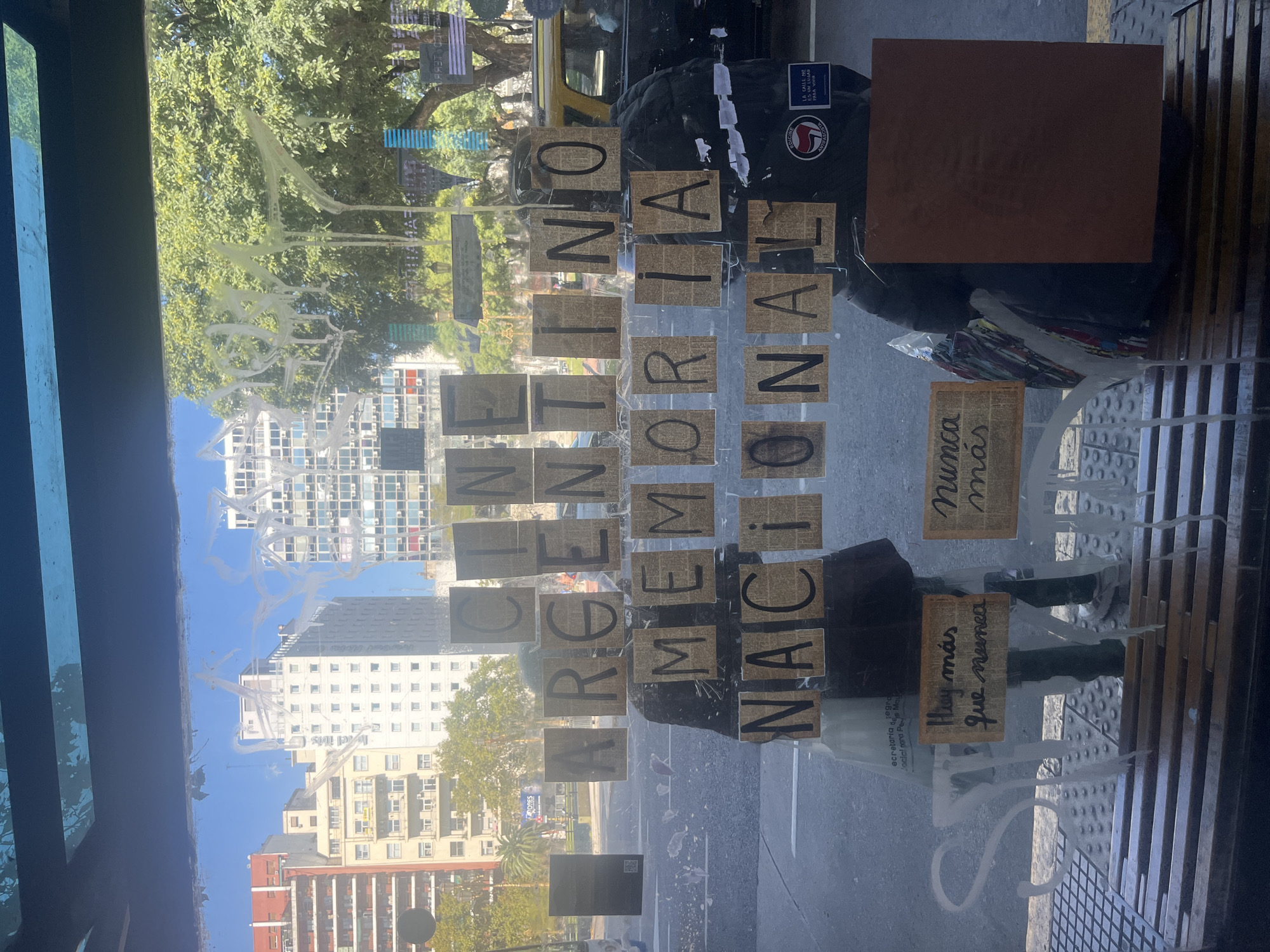 The image shows a storefront window display in Buenos Aires, Argentina. The words "CINE ARGENTINO MEMORIA NACIONAL" (Argentine Cinema National Memory) are displayed on paper cutouts taped to the glass. Below, additional paper signs read "Nunca Más" (Never Again) and "Hay más que nunca" (There is more than ever). A building and street scene are reflected in the glass, including the view of high-rise apartment buildings. The window is smeared with white residue. A trash bag and brown square-shaped object are visible in the upper right corner of the image. A sticker with the words "La Calle No es un lugar para vivir" (The Street is not a place to live) and a logo with the word "Antifascista" (Anti-fascist) are visible on the trash bag.