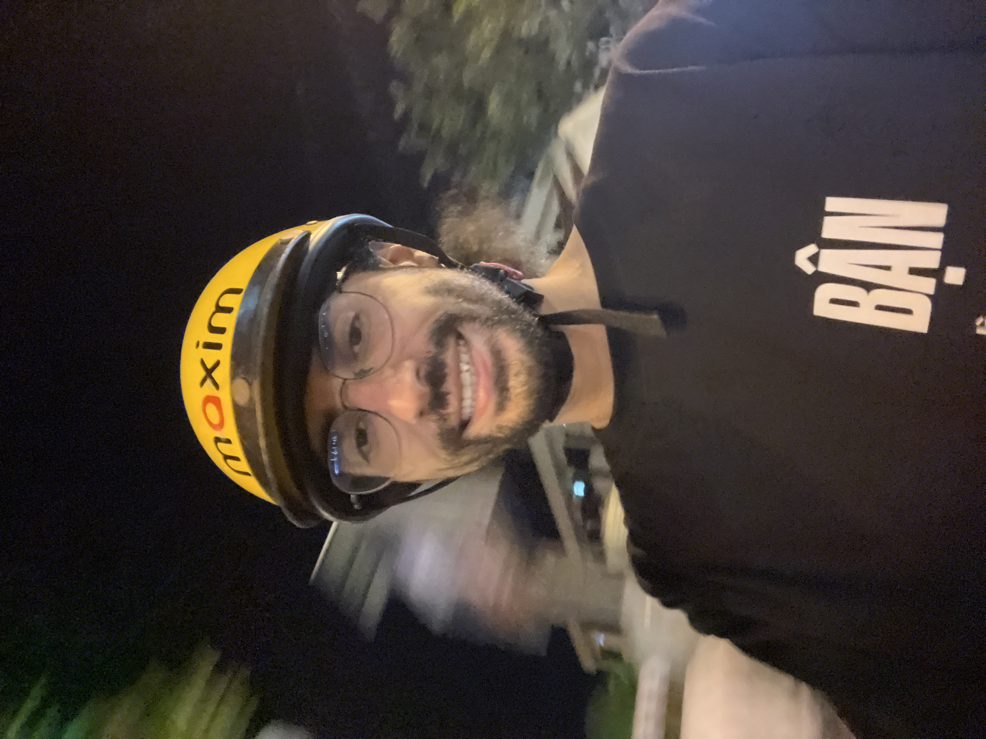 The image shows a person wearing a yellow helmet with "maxim" written on it in black. The person is also wearing glasses and a black t-shirt with "BÂn" written in white text. The person is smiling and appears to be outdoors at night, with some blurry foliage and lights in the background. The overall impression is that of a selfie taken by someone on the go, possibly a delivery driver. The location is identified as Nha Trang Ward, Vietnam. The image is rotated 90 degrees counter-clockwise.