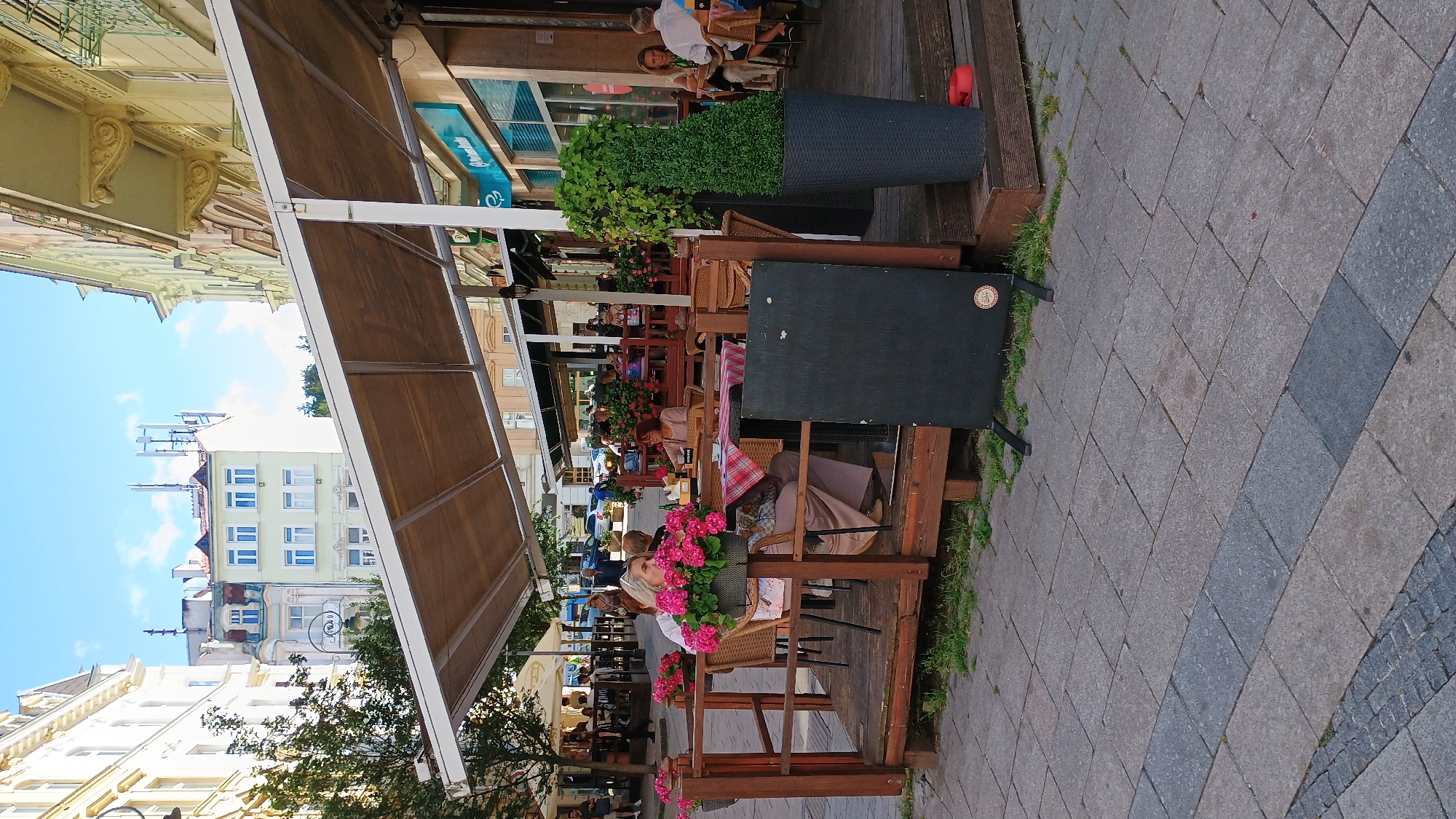 The image captures an outdoor seating area of a restaurant on a sunny day, likely in the city of Karlovy Vary, Czechia, given the architectural style. In the foreground, a wooden deck is set up with tables and chairs. Pink flowers in pots adorn the railing of the deck. A woman with blonde hair is seated at one of the tables, looking away from the camera. Another woman with red hair is seated at a nearby table, with a drink in front of her. A dark blackboard, possibly a menu board, stands on an easel on the deck. Behind the deck, a row of buildings with various architectural features can be seen, and further back, there are more buildings and trees. The ground is paved with cobblestones. The overall scene depicts a relaxed outdoor dining atmosphere in a European city.