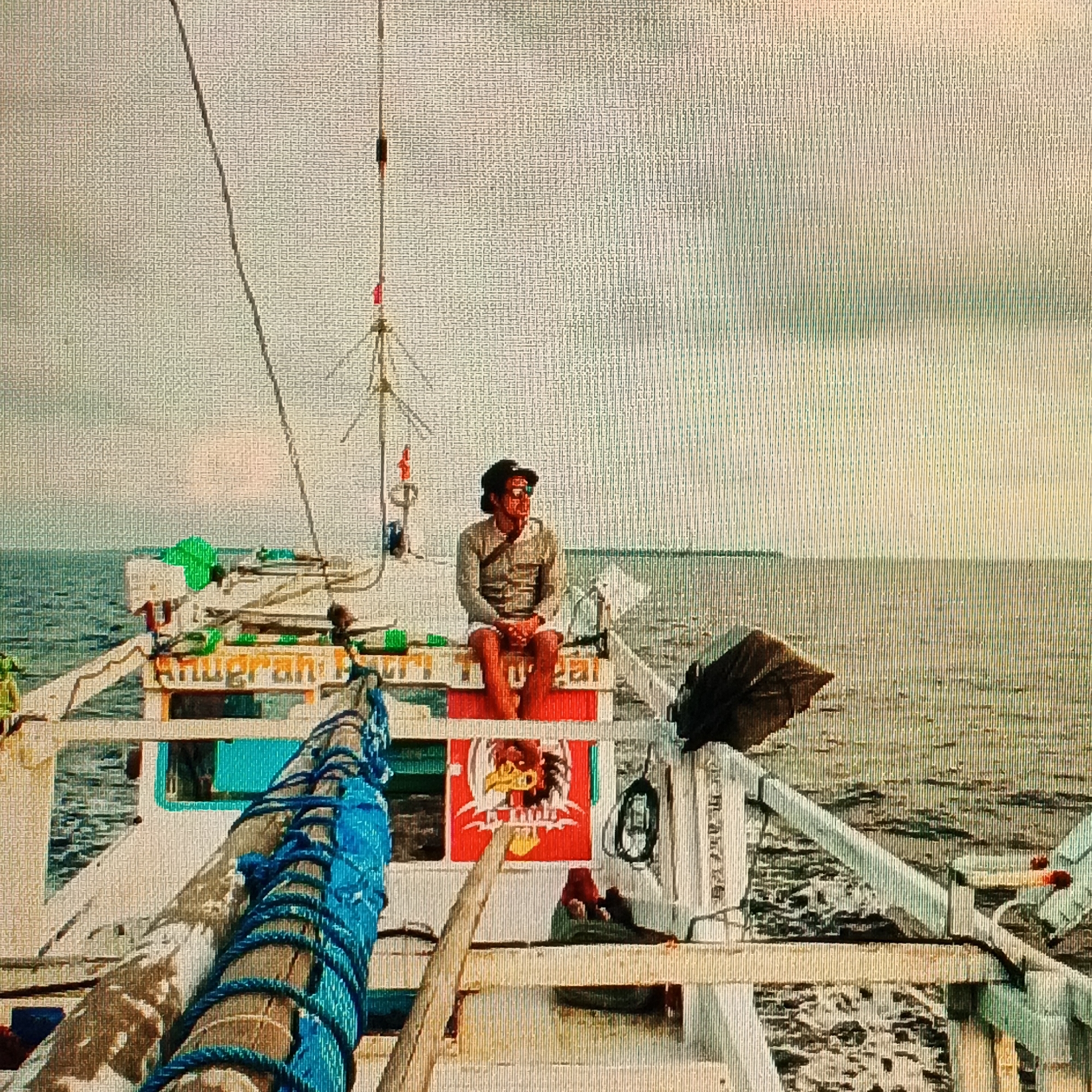 The image shows a man sitting on a boat on the ocean. The man is wearing a hat and a shirt. The boat is white and has a blue rope on it. There is a sign on the boat that says "Anugerah". The ocean is calm and there are clouds in the sky. The location is in the waters near Ende, Indonesia. The image appears to have been taken from another boat.