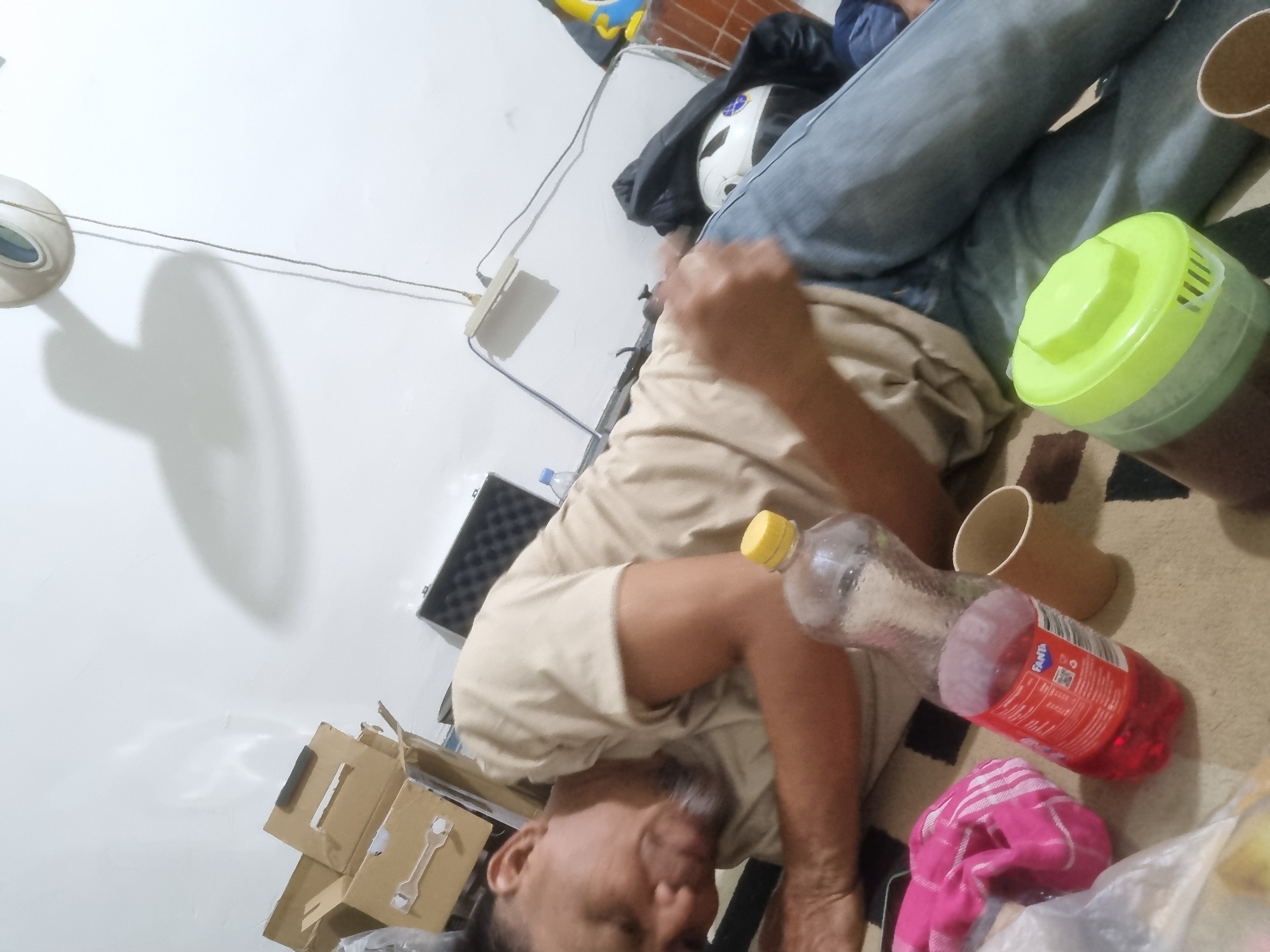 A man in a tan shirt and blue jeans is lying on his side on a carpeted floor. He appears to be sleeping or resting. In front of him are several objects, including a bottle of red Fanta, a brown paper cup, and a pink cloth. Behind him, there are cardboard boxes and a black case. The room appears to be dimly lit, with a shadow cast on the wall from a ceiling fan. The scene suggests a casual or informal setting, possibly a home or a storage area.