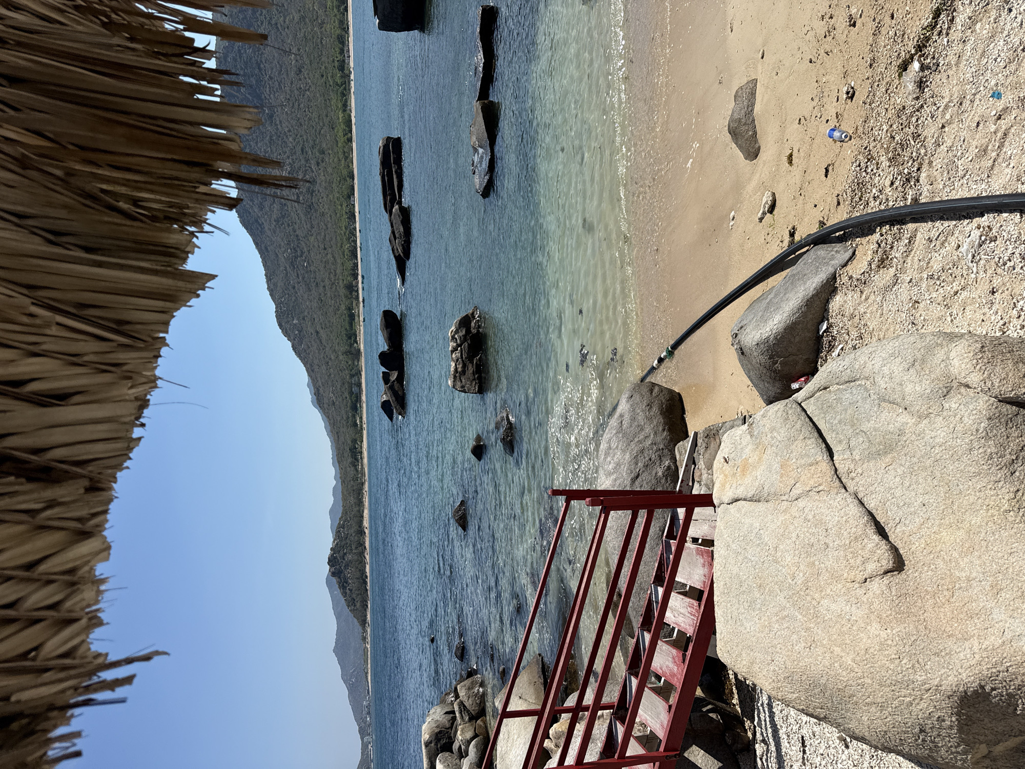 A view from Vinh Hai, Vietnam, displays a section of coastline. The composition features a thatched roof fragment in the upper left corner. The sea is dotted with dark-colored rocks extending from the coastline out into the water. A small, red metal staircase leads down towards the sea from the rocky shore. The beach is comprised of both sand and rocky terrain, with a discarded plastic bottle lying on the sandy part. A thick, black pipe stretches across the beach. The sky is clear and blue, and a mountainous landscape runs parallel to the coast.