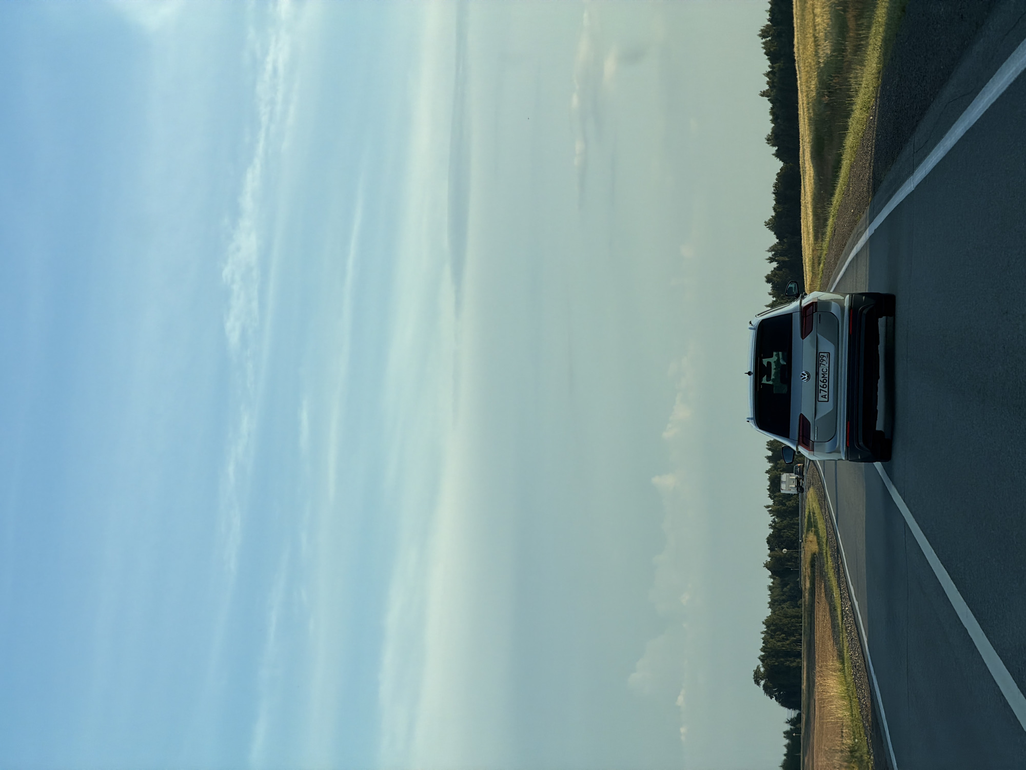 The image shows a silver Volkswagen SUV driving on a paved road in a rural setting. The road has a white line marking its edge and extends into the distance. The SUV is the primary object in the scene, viewed from the rear. The setting is characterized by a vast, pale blue sky with wispy white clouds, suggesting it's daytime, likely late afternoon or early evening due to the soft lighting. To the right of the road, there's a field of dry grass or crops, bordered by a dense line of dark green trees. The overall atmosphere is calm and serene, with no visible people or indication of immediate activity beyond driving. The location is identified as Obukhovo, Russia. A license plate on the SUV is partially visible, with the text "A 766 MC 799".