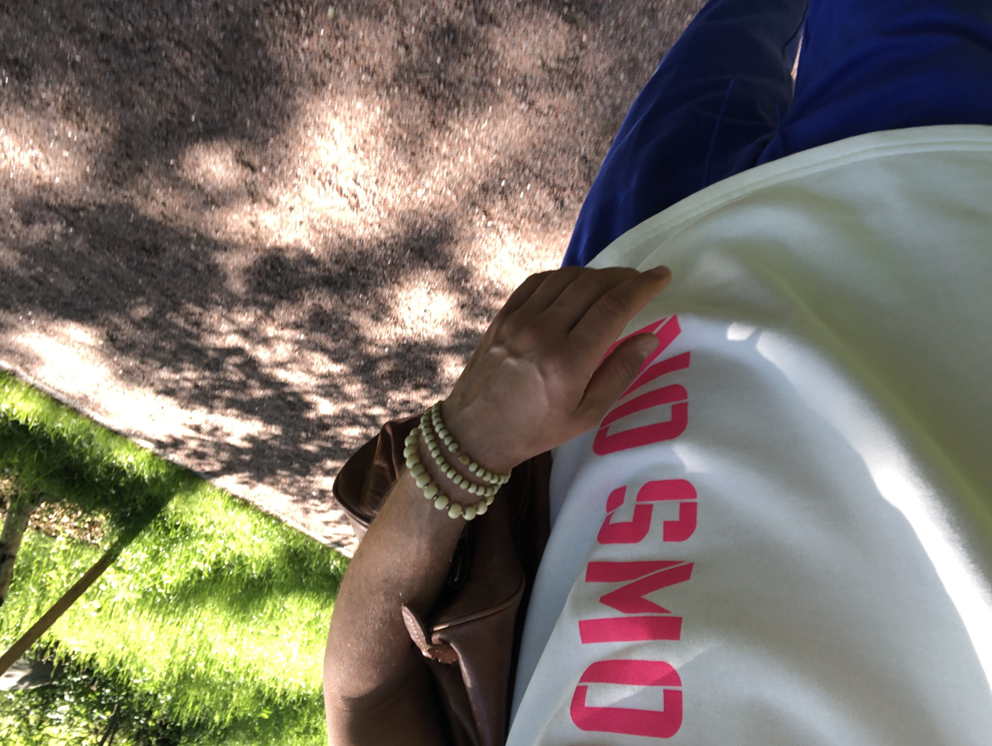 A person is walking on a dirt path outdoors, with grass and trees visible in the background. The person is wearing a white t-shirt with bright pink lettering that appears to read "OMG ON," blue pants, and a brown bag slung over their shoulder. They have three bracelets made of light-colored beads on their left wrist. The lighting suggests it is a sunny day, with dappled shadows on the path from the overhead foliage. The exact location in Saint Petersburg, Russia, cannot be determined from the image.