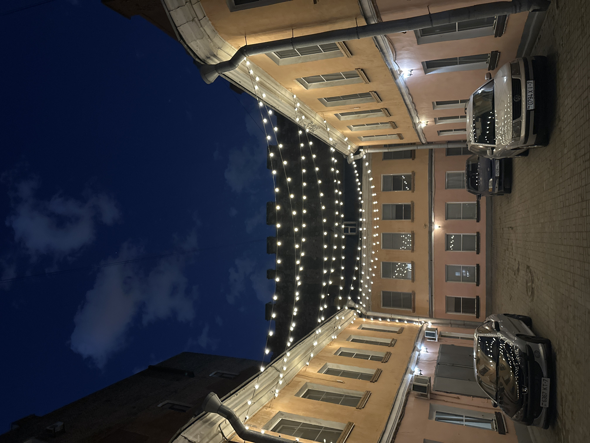The image depicts a courtyard in Saint Petersburg, Russia, at dusk or night. The sky above is dark blue, with some scattered clouds. The courtyard is surrounded by buildings with light-colored facades and rectangular windows. Strings of lights are suspended between the buildings, creating a festive atmosphere. Two cars are parked on the paved ground of the courtyard. The photograph has been rotated 90 degrees counter-clockwise.