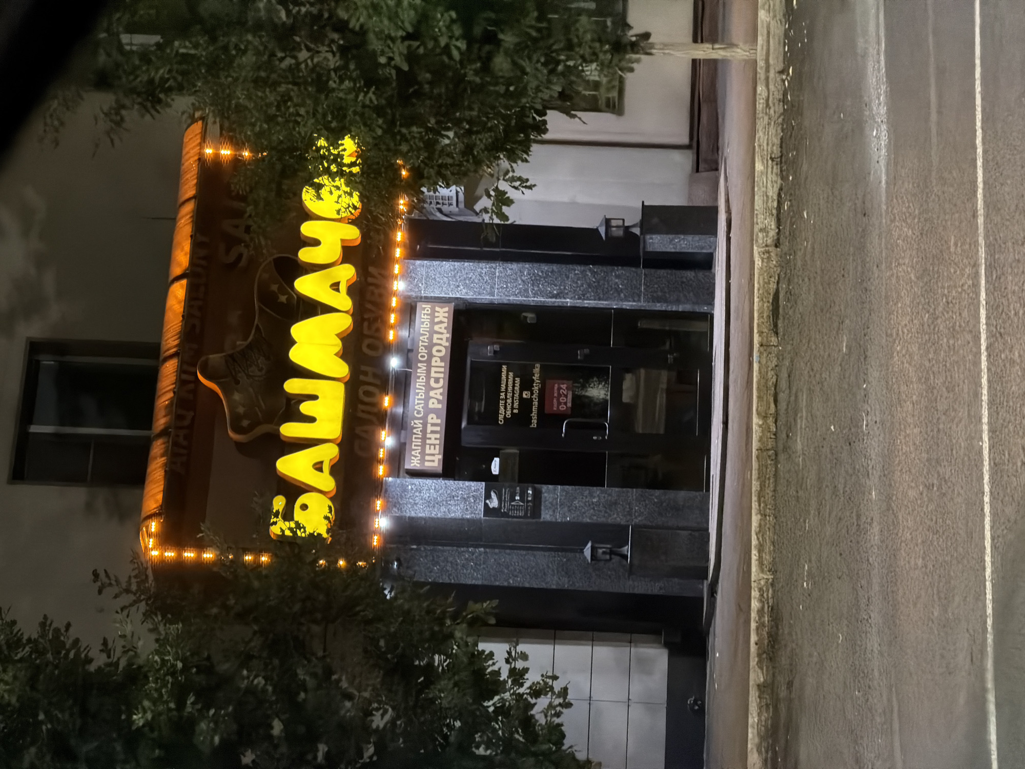 The image shows the exterior of a shoe store at night, likely in Astana, Kazakhstan, based on the Cyrillic text. The store has a brightly lit sign with yellow lettering that reads "БАШМАЧОК" and below it, "САЛОН ОБУВИ". There is also a smaller sign in purple text that says "ЖАППАЙ САТЫЛЫМ ОРТАЛЫҒЫ ЦЕНТР РАСПРОДАЖ". The entrance to the store is visible, with a glass door that has some informational text and a 0-24 hour indicator. The store appears to be on a street, with trees on either side of the entrance. The ground in front of the store is paved, and the street in the foreground is wet, suggesting it may have recently rained or is a rainy night. The lighting is primarily from the store's sign and some streetlights. There are no people visible in the image.