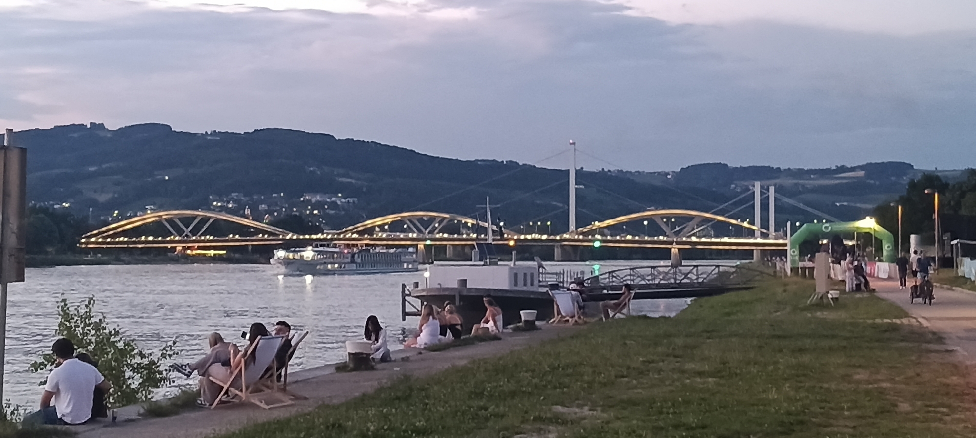 This image depicts a scene along the Danube River in Linz, Austria, during twilight. In the foreground, several people are seated on the riverbank, some in lounge chairs, enjoying the view. A man in a white t-shirt is seated on the ground, facing away from the camera. Further along the bank, a group of people are gathered, some sitting and some standing, near a large green inflatable archway.
The river occupies the middle ground, with a large passenger ship sailing across it. In the background, a series of illuminated bridges span the river, with a suspension bridge visible further in the distance. The bridges are brightly lit with yellow lights, creating a striking contrast against the darkening sky and the silhouetted, tree-covered hills. The overall atmosphere is serene, with the soft evening light creating a calm and inviting ambiance. The setting appears to be a public area, possibly a promenade or park, given the presence of seating and the archway, which might indicate an event or a starting/finishing line for an activity.