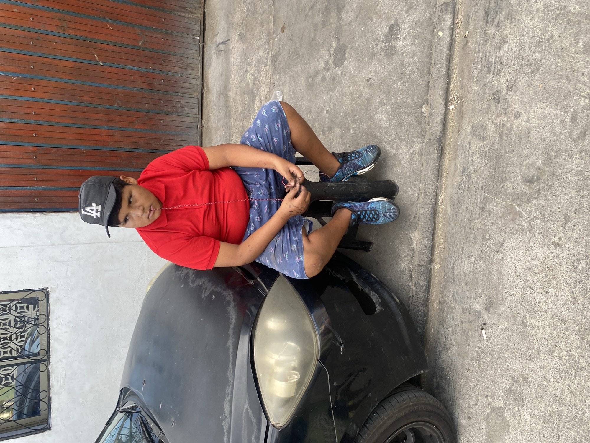 A medium shot, taken in daylight, shows a young boy, approximately 10-12 years old, sitting cross-legged on the hood of a black car. The boy wears a red t-shirt, patterned blue shorts, blue athletic shoes and an LA baseball cap. He faces the camera, slightly turned left, holding and manipulating a length of red wire in his hands. A black tire rests under his crossed legs. The car appears to be an older model, with visible wear and tear to the paint. The background includes a wooden panel wall to the left and a concrete surface.
Based on architectural features and available knowledge, the location is possibly a residential or light industrial area in Arica, Chile. Concrete and wood construction, similar to the materials shown in the photograph, are typical in this region.