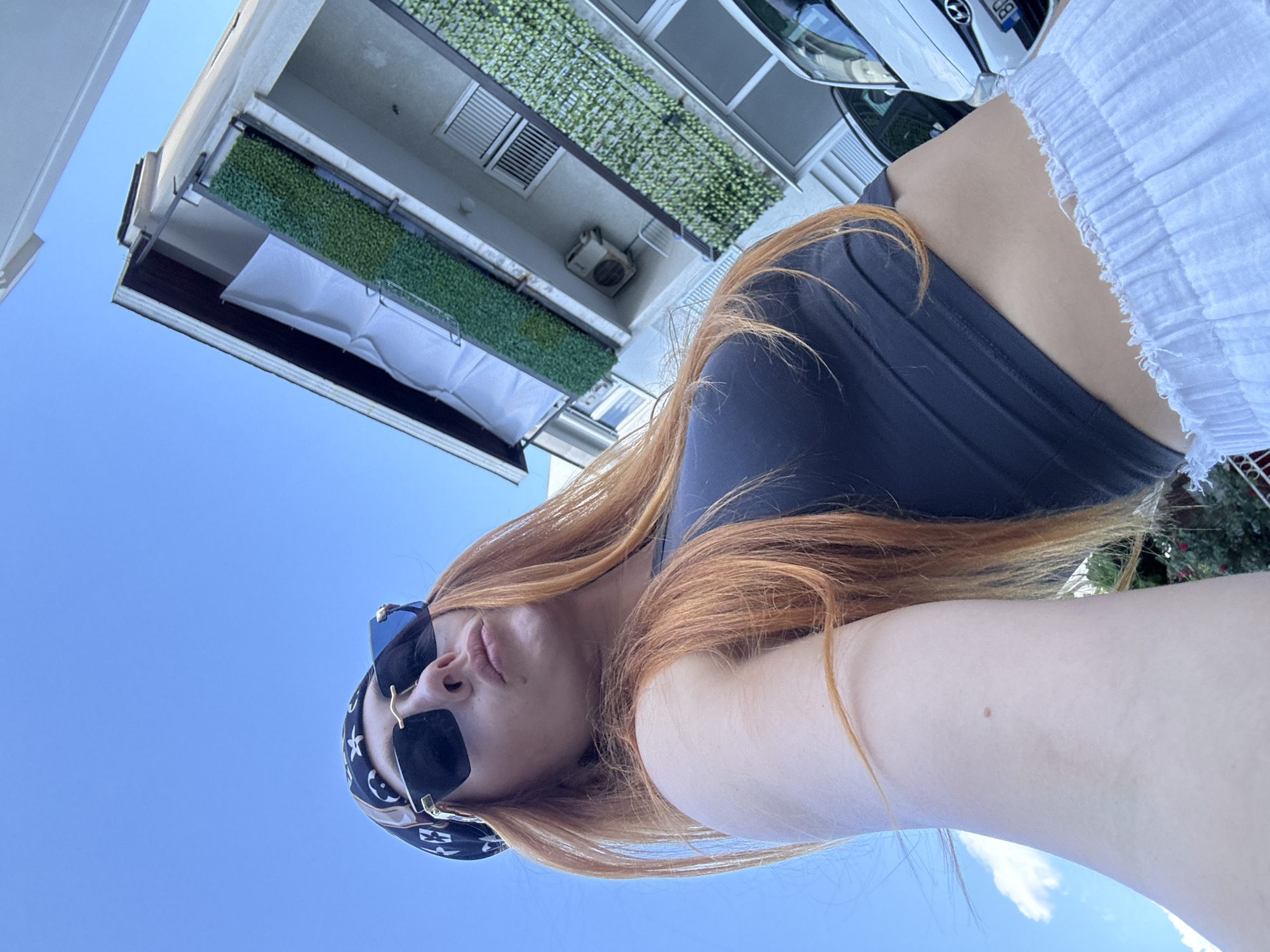 A young woman with long, reddish-blonde hair is taking a selfie outdoors. She is wearing a dark, strappy top and white, ruffled shorts. She has on dark sunglasses with a gold frame and a black bandana with a white pattern around her head. Her mouth is pursed, as if she is about to kiss. The background consists of a bright blue sky, the side of a building with white siding and balconies adorned with artificial greenery, and an air conditioning unit. A white car is partially visible. The image appears to have been taken during the daytime, on a clear day, likely in a warm climate. The angle of the shot suggests the woman is standing and holding the camera above her head, looking up at the sky. The presence of the building and car indicates an urban or suburban setting, possibly a residential area.