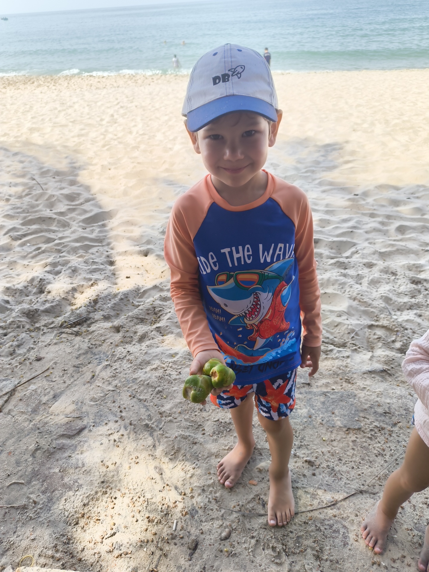 In Phu Thuy Ward, Vietnam, a young boy stands on a sandy beach, wearing a blue and white baseball cap and an orange and blue rash guard with a shark graphic. He is holding a small cluster of green fruits in his right hand. The beach has rippled sand and scattered debris, with the ocean visible in the background. Another child's bare legs are visible on the right side of the frame.