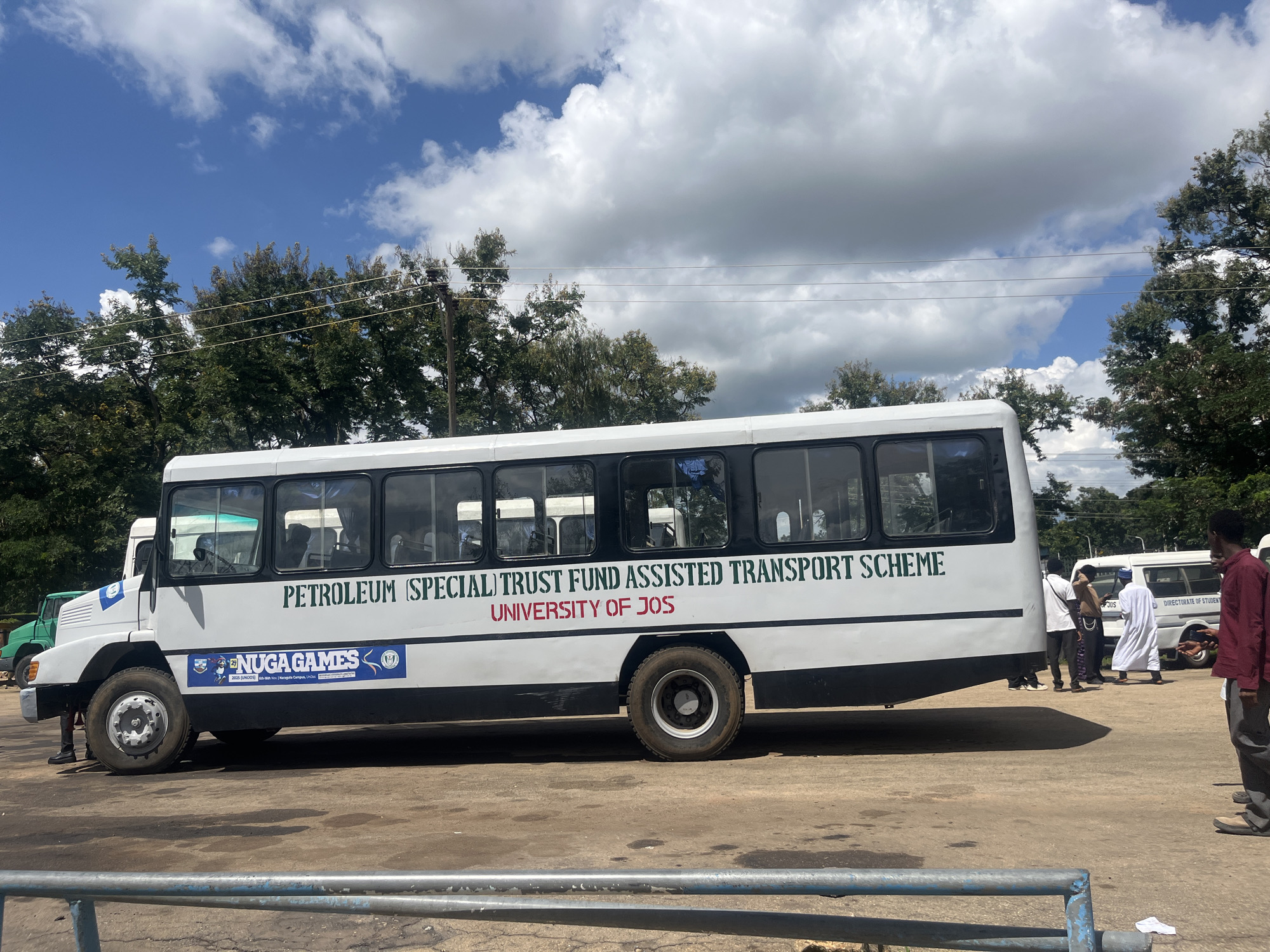 The image shows a white bus with "PETROLEUM (SPECIAL) TRUST FUND ASSISTED TRANSPORT SCHEME" and "UNIVERSITY OF JOS" printed on its side. A blue banner with "NUGA GAMES 2025 (UNIDOS)" is also visible on the bus. Several people are standing near the bus, some of whom appear to be students. A green truck is parked behind the bus. The scene takes place outdoors on a sunny day with scattered clouds. The ground is paved with dirt and some puddles. The presence of "UNIVERSITY OF JOS" and "NUGA GAMES" suggests the location is likely related to the university in Jos, Nigeria. The overall atmosphere appears to be casual and perhaps a gathering or event is about to commence or has recently concluded.