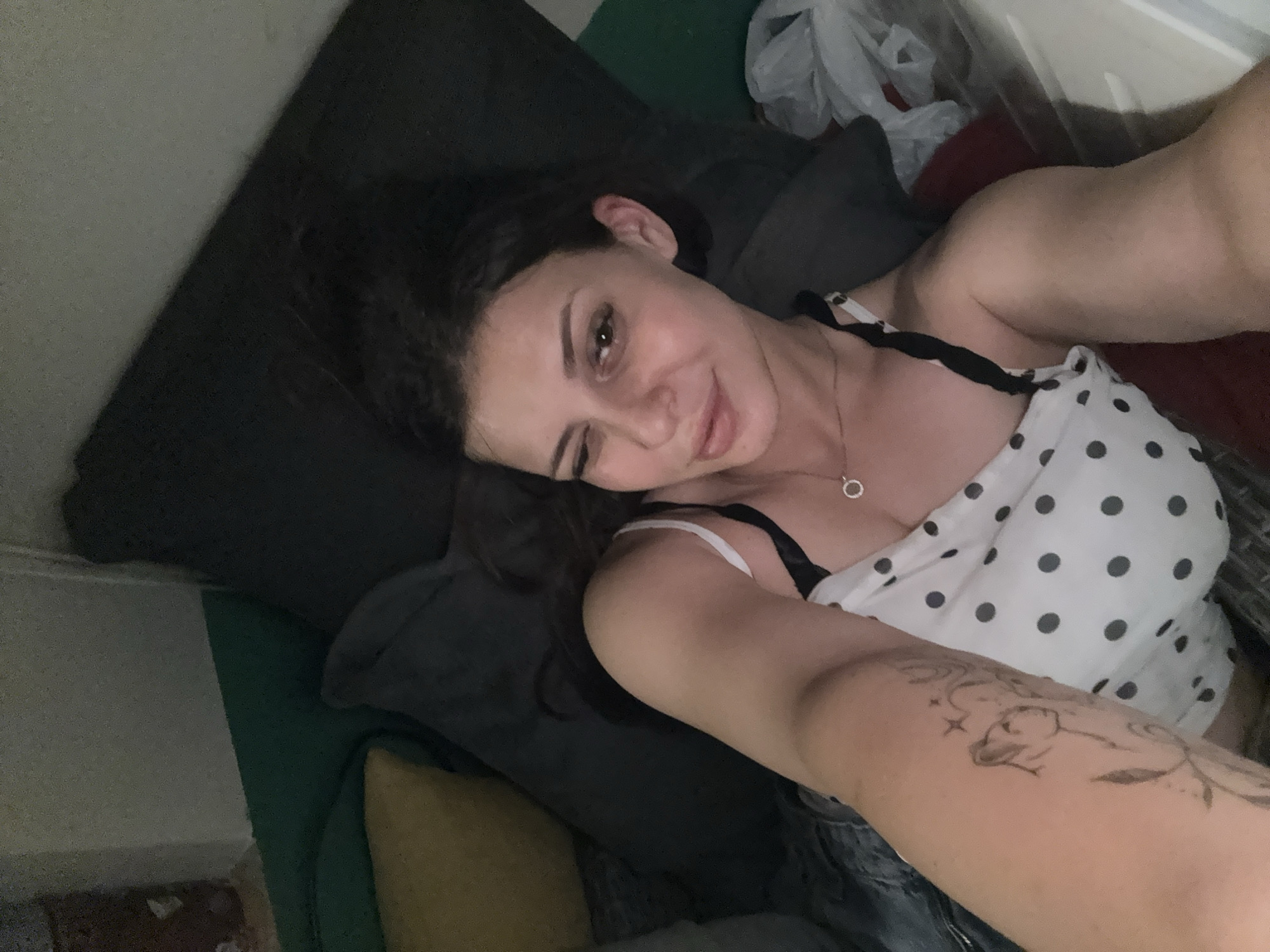 The image shows a woman lying on a couch. She is wearing a white polka dot top with black straps and a thin silver necklace. She has dark hair and is winking at the camera. Her left arm is visible, and it has a tattoo of a woman's face and some swirls. The couch appears to be in a dimly lit room, possibly a living room or bedroom. There are several pillows on the couch, some dark gray and one yellow. To the right of the woman, there is a white plastic bag. The image does not provide any information about the location in the city of Plovdiv, Bulgaria.
