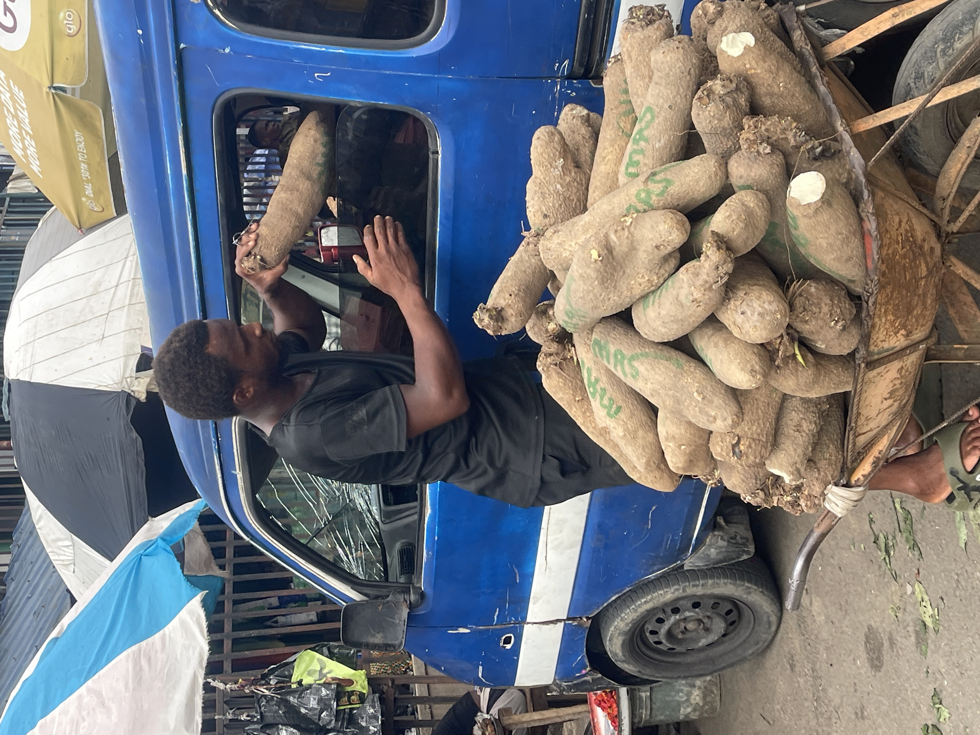The image, correctly oriented, captures a vibrant outdoor market or street scene, likely in a West African setting. A man, wearing a black t-shirt, is seen in the driver's seat of a blue minibus or van, holding a large, elongated yam. He appears to be either loading or unloading the yam through the open driver's side door. Another person is faintly reflected in the vehicle's side window.
The blue vehicle, distinguished by a white stripe along its side, has a visibly cracked windshield and a front passenger tire in view. It is heavily loaded with numerous large yams, which are piled high on a wooden cart or frame secured to its side, extending from the vehicle. Some of the yams have green markings, with "MAS" visible on several. A bare foot, clad in a camouflage-patterned sandal, is visible near the yam cart on the right side of the frame.
The background features several umbrellas, including a prominent yellow one with the "Glo" logo and the text "MORE DATA MORE VALUE" and "DIAL *108*1# to enjoy." Other umbrellas are white and black, and blue and white. Scattered leaves are visible on the ground, and various structures made of corrugated metal and other materials suggest a bustling commercial environment. The scene is illuminated by daylight.