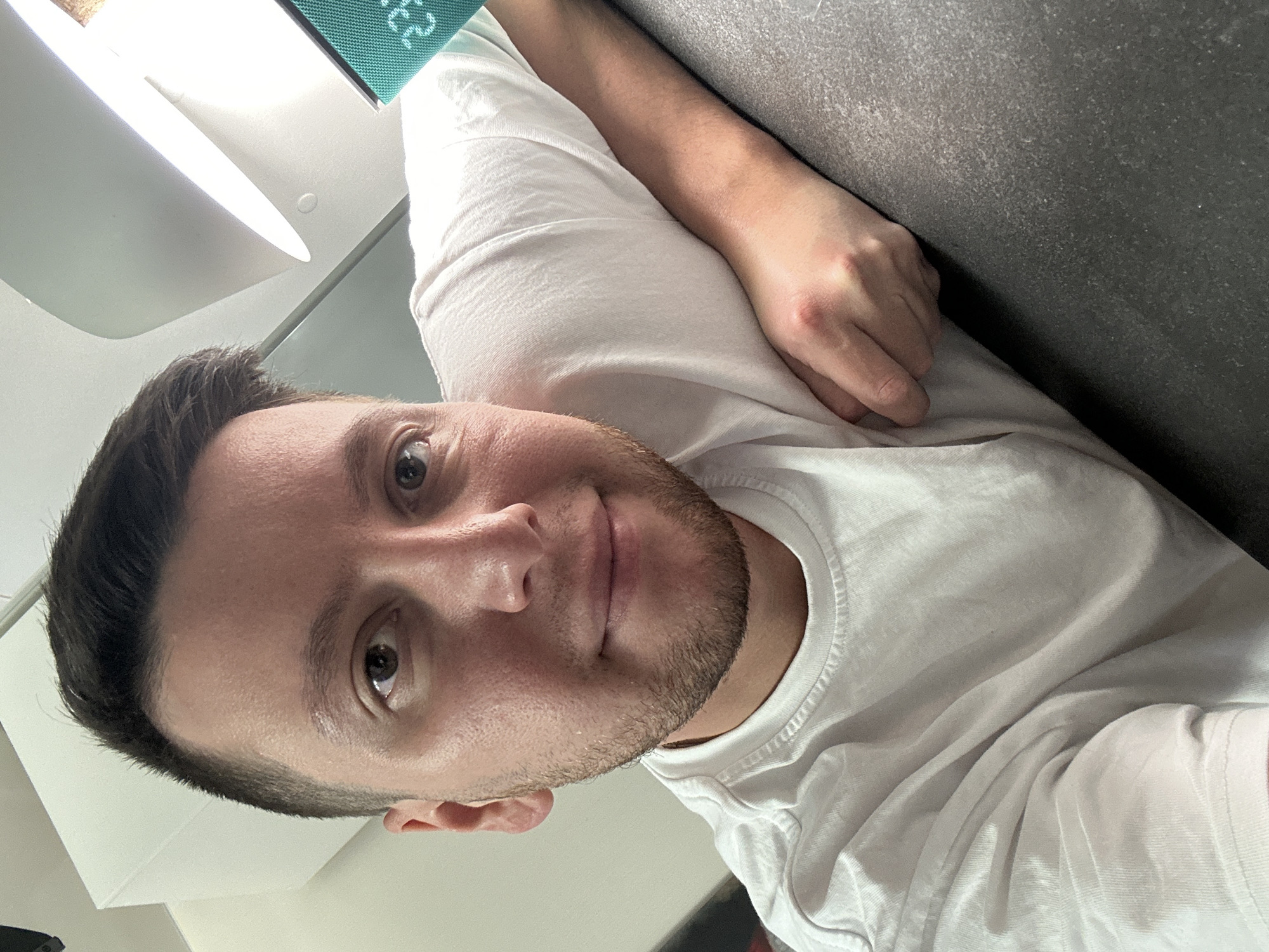 A close-up, canted, upward-facing portrait depicts a man with fair skin, a short, dark-brown haircut, and a stubbly beard, wearing a white t-shirt. His arm rests on a dark-grey surface. In the background, part of a white object and a turquoise item are visible. There is no information to confirm that this is the city of Мирный, Russia.