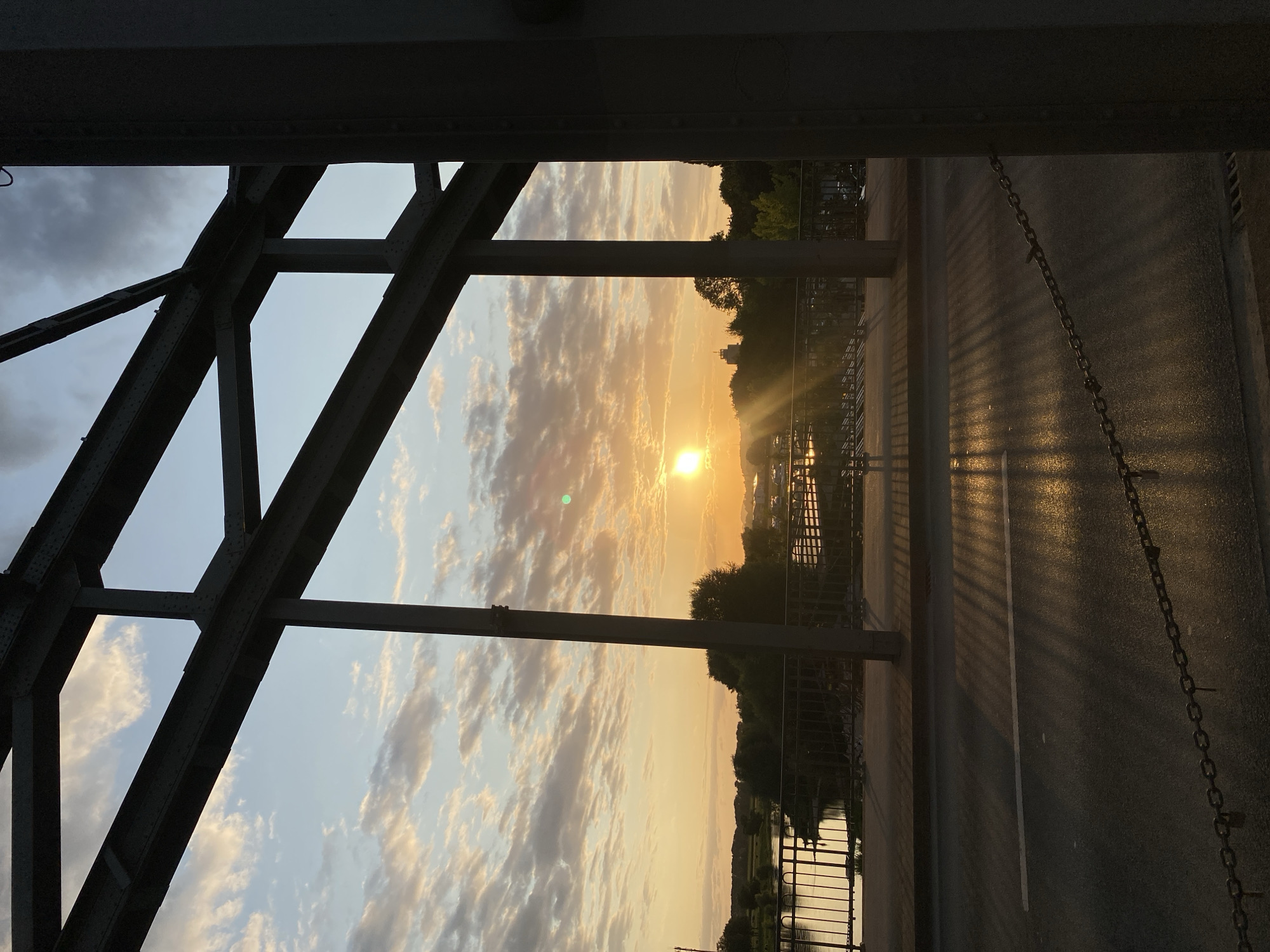 The image captures a sunset viewed from a bridge in Rinteln, Germany. The perspective is from the road deck of a metal truss bridge, looking towards the horizon. The sun, a bright orb with visible rays, is low in the sky, casting a warm, golden light across the scene. The sky is a mixture of pale blue and scattered clouds, tinged with orange and yellow hues from the setting sun.
Below the bridge, a body of water, likely the Weser River, reflects the sky's colors. A metal railing runs along the edge of the bridge overlooking the river. On the far bank, a treeline with varied foliage is visible, interspersed with some buildings and a prominent tower structure. The road on the bridge is asphalt, with a white line marking a lane. The sunset's light creates long, striped shadows on the road, cast by the bridge's structure. A heavy metal chain runs along the side of the road on the bridge deck. The overall atmosphere is serene and picturesque, highlighting the natural beauty of the sunset framed by the industrial structure of the bridge.