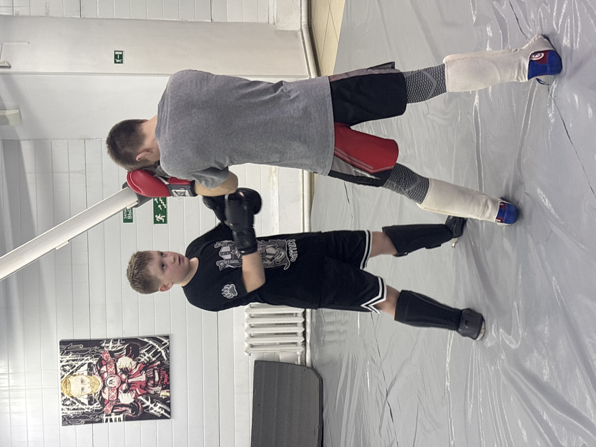 Two young males are practicing martial arts in an indoor training facility. The boy in the foreground is wearing a black t-shirt, black shorts with white stripes, black shin guards, and black gloves, and is in a combat stance. The boy in the background is wearing a grey t-shirt, black shorts, white shin guards, red boxing gloves, and is holding a red padded target. A painting of a man with a beard and an ornate red and white outfit hangs on the white wall. A radiator is against the wall near the first boy. The floor is covered in a large, thin sheet of grey plastic. The location is likely in the city of Perm, Russia, given the presence of Cyrillic text.