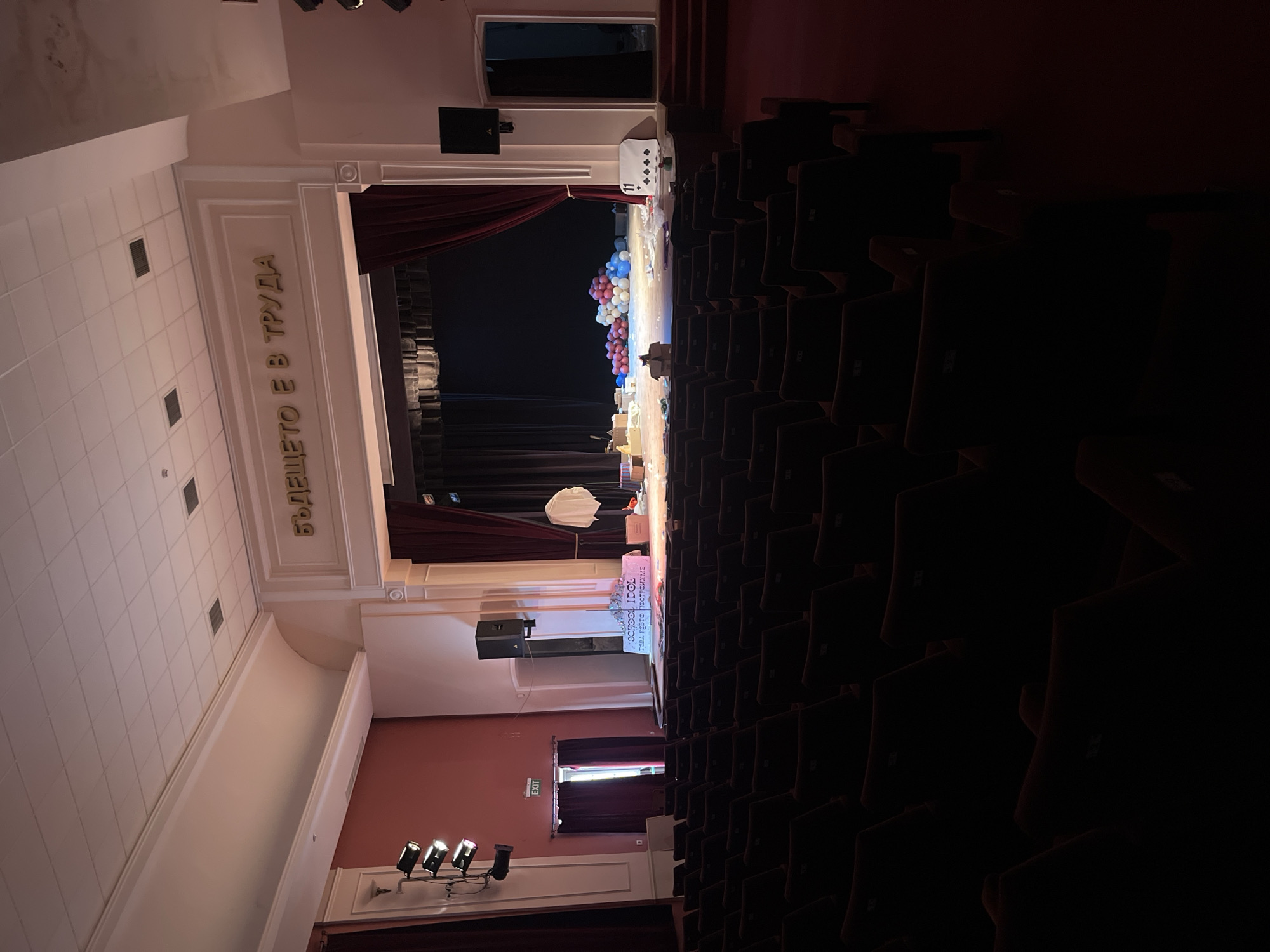 The image shows the interior of a theater in Nova Zagora, Bulgaria. The theater is equipped with rows of seats, a stage with a dark backdrop, and red curtains on the sides. On the stage, there is a cluster of balloons and a white structure that could be part of a stage setup or decoration. The theater's name or a related phrase is inscribed in a script above the stage. The lighting is uneven, with the stage and some sections of the theater more illuminated than others. There is an exit sign visible on the left side of the image. The scene appears to be set up for an event.