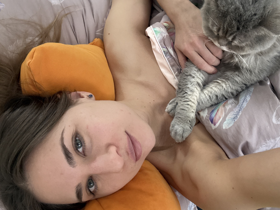 An overhead photograph depicts a single adult female and a domestic cat in a bed, located in Saint Petersburg, Russia. The female, positioned on her back with her head resting on two orange pillows, has light skin, dark brown hair, and blue-green eyes gazing directly forward. Her lips are slightly parted. She exhibits minimal makeup. Small, dark stud earrings are visible in her ears. Her bare shoulders and upper chest are exposed above a light-colored duvet featuring muted feather patterns and a pink trim. Her right arm is bent, extending out of frame, suggesting she is holding the camera. Her left hand is partially visible, resting on the duvet near the cat. A grey-furred cat is situated to the right of the female, partially under the duvet, with its head turned away from the camera. Its left front paw is visible resting on the duvet. The setting appears to be an indoor bedroom, illuminated by natural light.