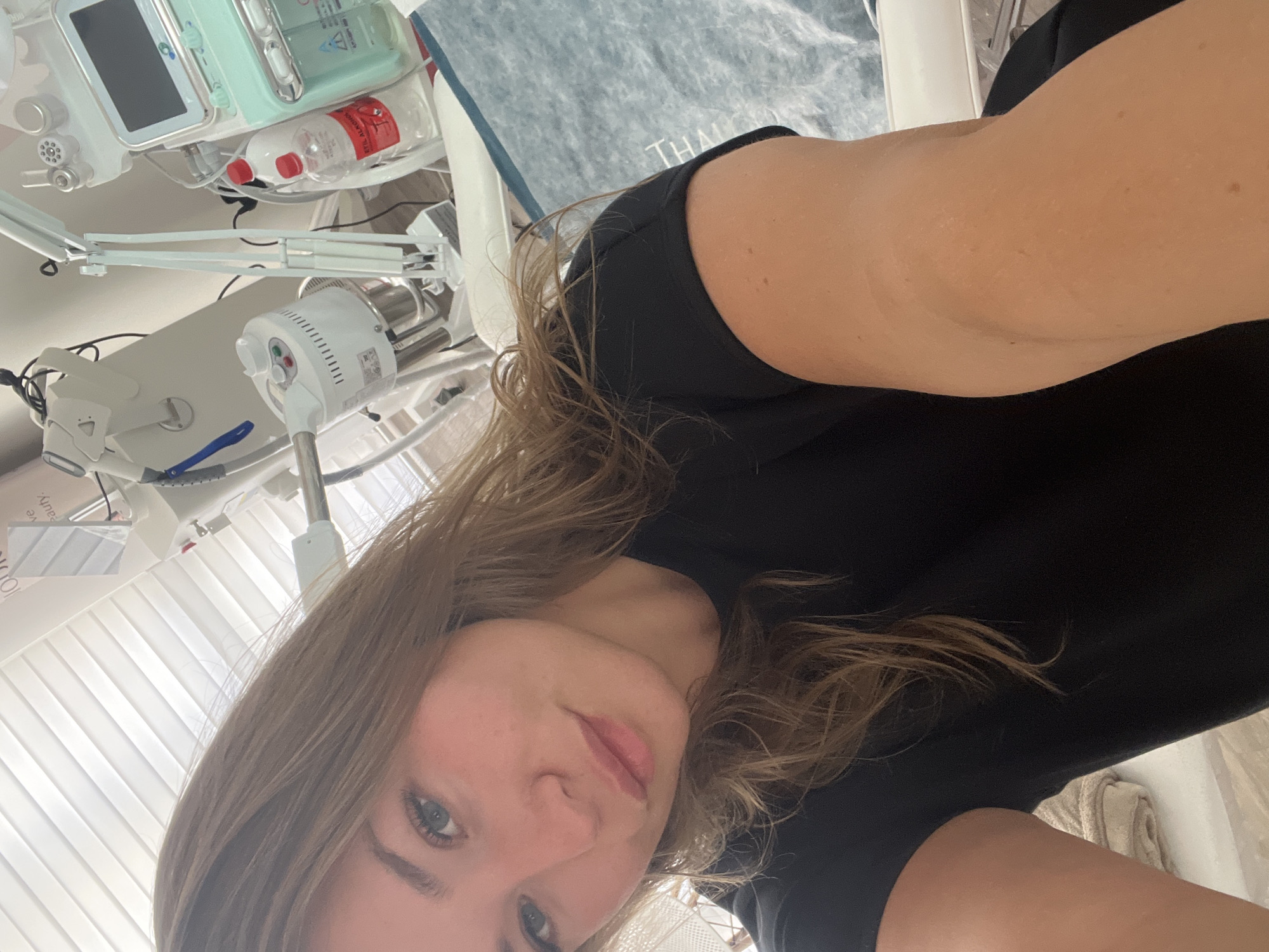 A selfie of a fair-skinned woman with light brown hair, wearing a black shirt, taken in what appears to be a medical or spa setting. The background includes medical equipment such as a facial steamer, a machine with a screen, and bottles of liquids. The location is possibly a spa or clinic in Budva, Montenegro.