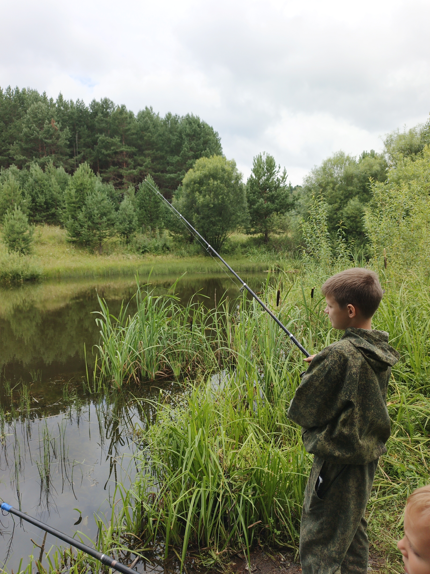 A boy in camouflage clothing stands by a pond in Medvedyovka, Russia, holding a fishing rod. The pond is surrounded by tall green grass and reeds, with a forest in the background. The sky is overcast. Another child's head is partially visible in the lower right corner of the image.