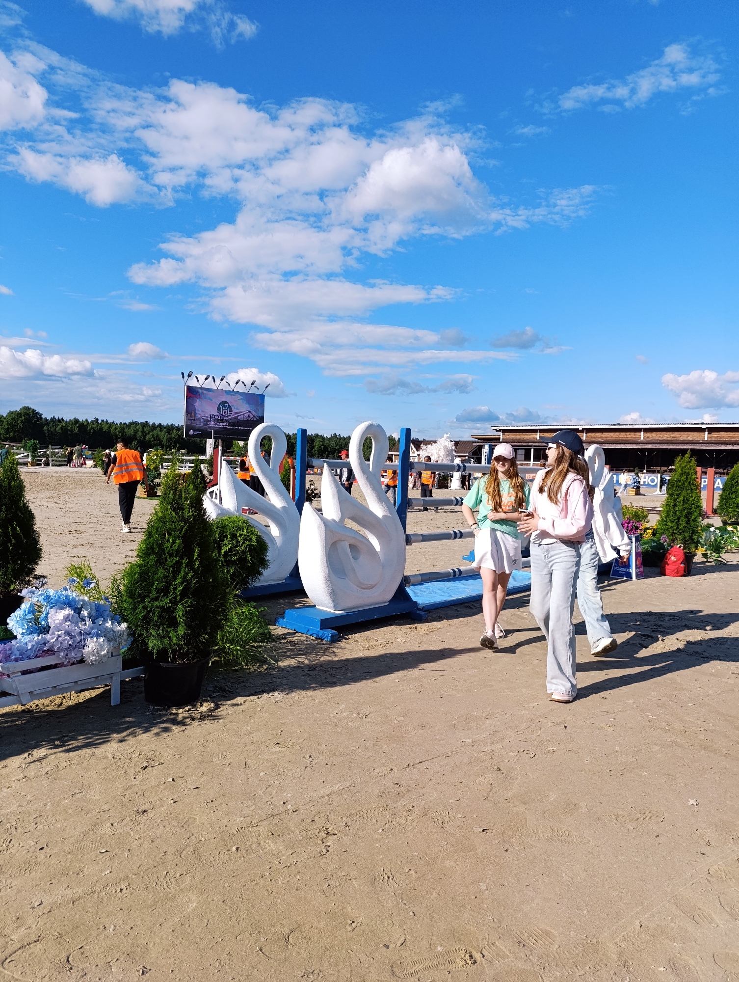This media file shows an outdoor scene with several people, decorative objects, and a sandy ground typical of an equestrian event. In the foreground, three young women are walking. Two of them are wearing baseball caps, and one is wearing a light green t-shirt and white shorts, while the other is in a light pink hoodie and light blue jeans. Behind them, there are large white swan-shaped sculptures on blue bases, and further in the background, a large digital billboard displays event information.
A person wearing an orange vest is walking away from the camera towards the left side of the frame, suggesting they might be staff or an organizer. Several evergreen trees in potted planters are also visible, along with a decorative arrangement of blue and purple flowers in a white planter. The scene appears to be set during a daytime event, with clear blue skies and scattered white clouds, indicating fair weather.
In the background, there are wooden structures, possibly stables or event buildings, and fencing, which are common features at horse riding competitions. The sandy terrain suggests an arena or a pathway within such a venue. The overall atmosphere seems relaxed and possibly celebratory, given the presence of decorative elements and people enjoying the event. The billboard shows some text and logos, but they are not clearly legible. The location context is Ivanovskoe, Russia.