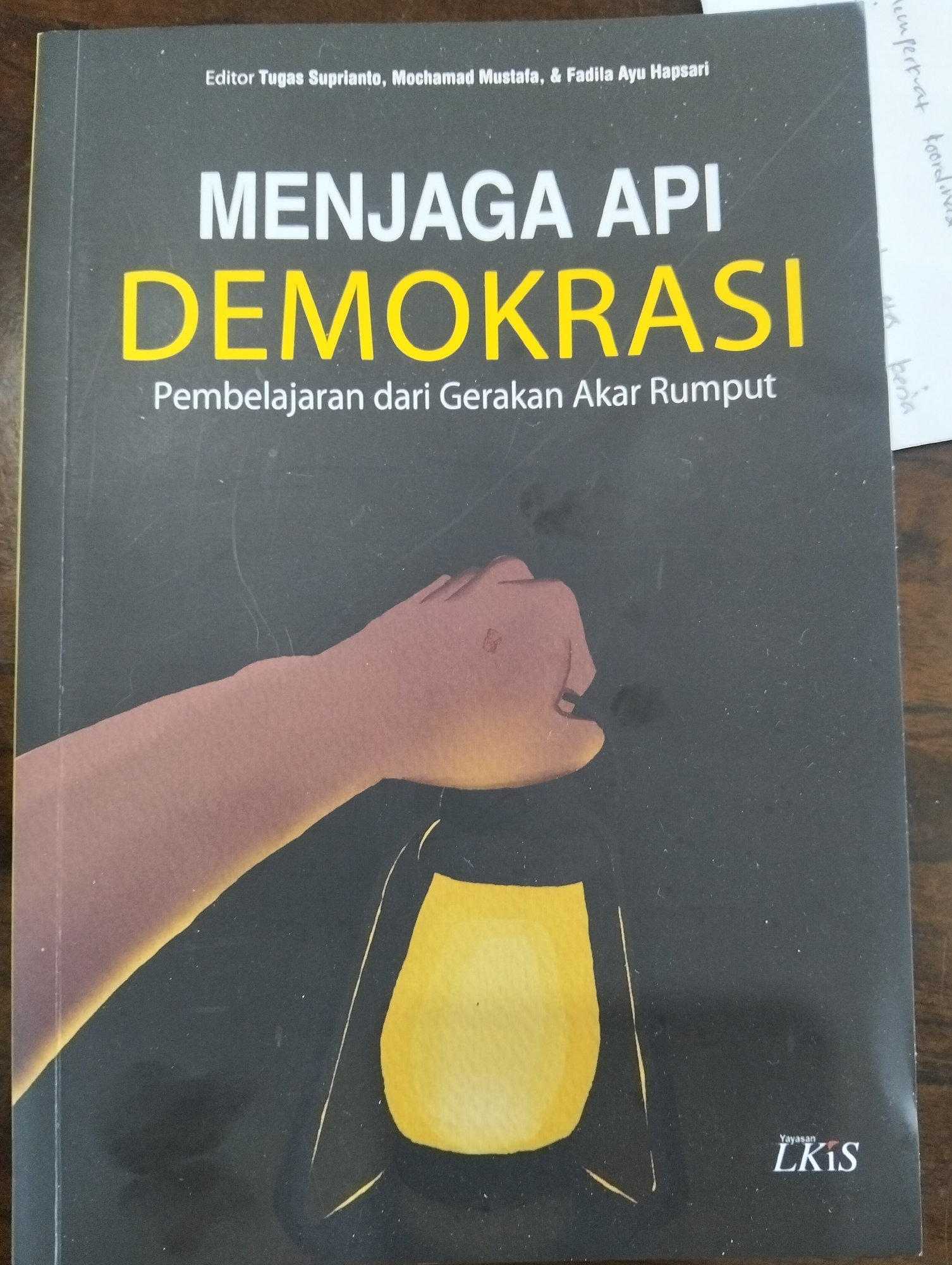The image displays the cover of a book titled "MENJAGA API DEMOKRASI: Pembelajaran dari Gerakan Akar Rumput" (Keeping the Flame of Democracy: Lessons from Grassroots Movements). The cover is predominantly dark gray with white and yellow text. At the top, the editors are listed as Tugas Suprianto, Mochamad Mustafa, & Fadila Ayu Hapsari. A graphic of a hand holding a lit lantern is positioned in the lower half of the cover, casting a warm yellow glow. The publisher's logo, "Yayasan LKIS," is visible in the bottom right corner. There is no visible connection or mention of the city of Julicotmeurak, Indonesia, within the image. The image itself is a close-up photograph of the book cover, likely taken indoors.