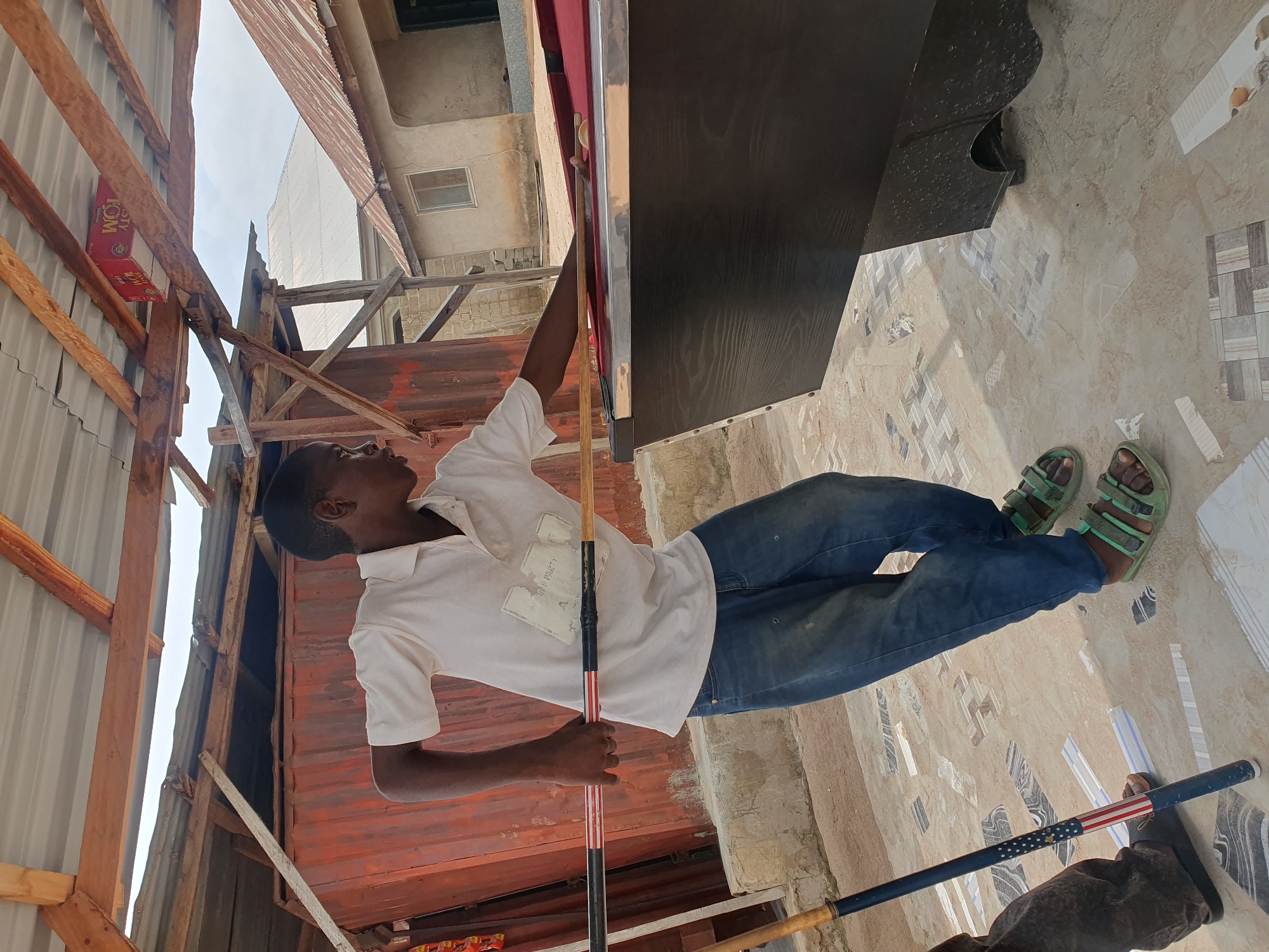 In Lokoja, Nigeria, a young male is playing pool at a makeshift outdoor billiards setup. He is wearing a white polo shirt and blue jeans, with green sandals on his feet. He holds a pool cue, aiming at a ball on the red felt-covered table. The table is positioned outdoors, under a corrugated metal roof supported by wooden beams, suggesting a humble, informal establishment. The background shows weathered buildings and a large red corrugated metal structure, typical of parts of Lokoja. The ground is paved with patterned tiles. Another person's legs are visible on the left, holding a pool cue decorated with an American flag motif, and wearing dark footwear. The scene appears to be a common leisure spot in Lokoja, Nigeria.