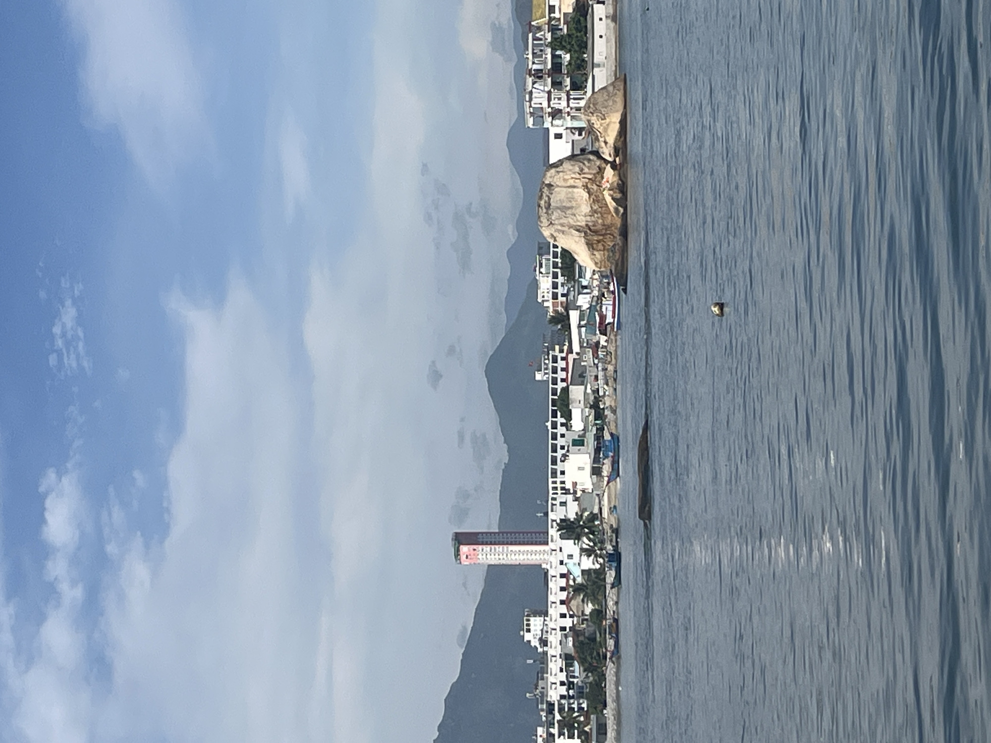This photograph, taken in Bac Nha Trang Ward, Vietnam, presents a coastal scene. On the left, a clear blue sky with scattered white clouds fills the frame. Moving right, a mountain range acts as a backdrop to a densely populated coastline. Buildings, predominantly white, extend along the shore, punctuated by palm trees. A tall, red and white building stands out among the others. In the water, a large rock formation sits near the shoreline. The sea, in varying shades of blue, occupies the right side of the frame, displaying wave patterns.