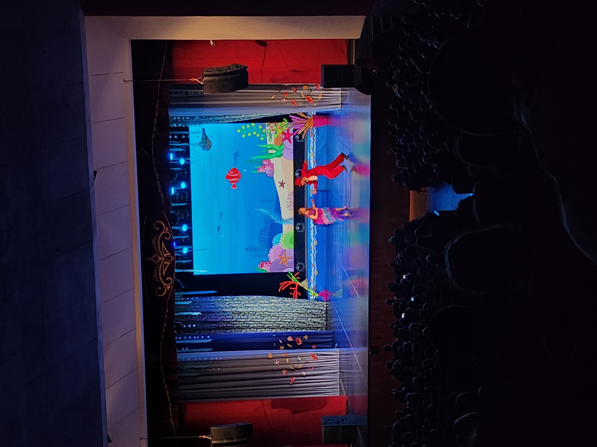 The image depicts a vibrant live stage performance in a theater, located in Chaikovsky, Russia. The stage is elaborately decorated with an underwater theme, featuring a bright blue backdrop adorned with colorful, cartoonish elements such as coral formations, green and pink seaweed, starfish, and various marine life, including a prominent red lantern-like fish and another grey fish.
Two performers are visible on the brightly lit stage, appearing to float or dance. One figure, dressed in a striking red suit with gold accents and a small hat, is positioned in the upper-mid section of the stage. The second performer is dressed in a multi-colored, predominantly purple and pink, flowing garment, located below the first. The stage floor reflects the blue and white lights, enhancing the aquatic ambiance.
The stage design incorporates multiple layered backdrops and props. Some layers feature textures resembling stone or fabric, while others display decorative patterns and golden swirls. The lowest visible stage layer is adorned with scattered objects resembling seashells and small marine creatures. To the right of the stage, a densely packed, dimly lit audience is visible, observing the performance. The overall scene suggests an engaging and lively production, likely a children's play or musical, taking place indoors, presumably in the evening. No visible text is present in the image.