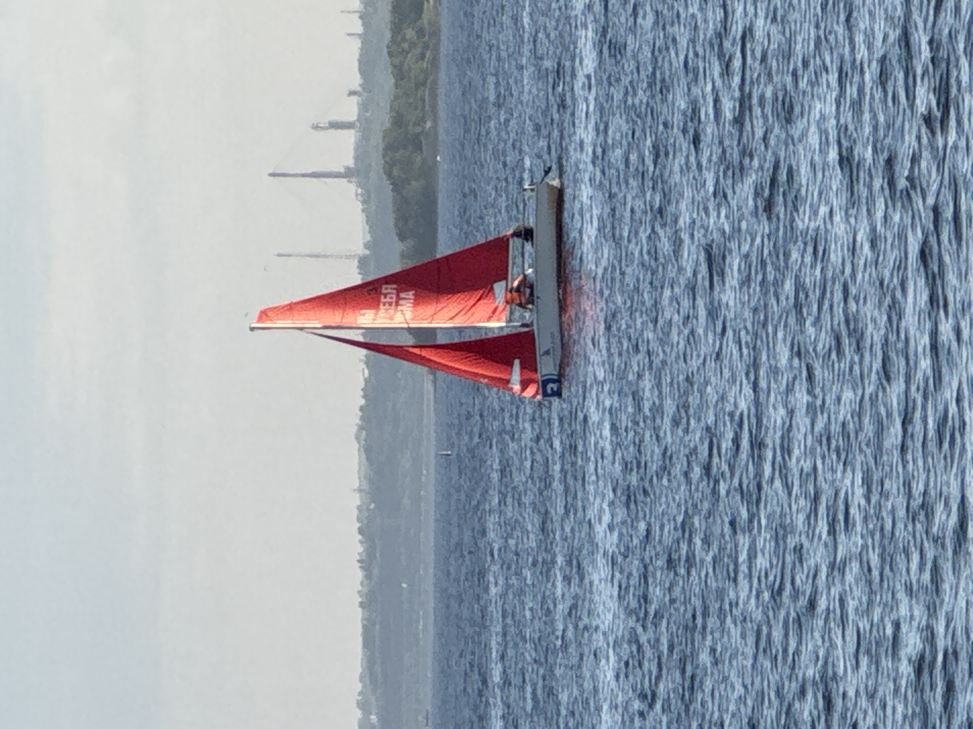 A sailboat with red sails is on the water. There are two people on the sailboat. The sailboat has writing on the sail. In the background, there is a city skyline with industrial structures. The sky is clear. The location is the Volga River in Saratov, Russia.