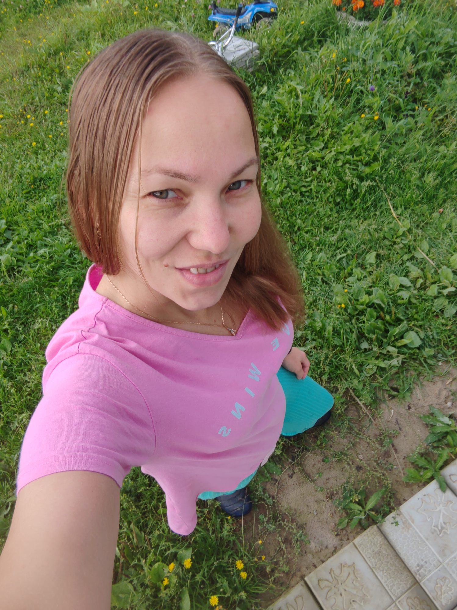 In this outdoor, high-angle shot taken in Чухлэм, Russia, a young woman is pictured from the chest up, looking up and smiling at the camera. She has shoulder-length brown hair that is slightly damp and parted in the middle. Her skin is fair, with a smattering of freckles and a mole on her right cheek. She is wearing a bright pink t-shirt with light blue lettering visible on the front. The text appears to read "I M I W" backward, suggesting it might be "WIM I" or a similar mirrored phrase. She also has a delicate gold chain necklace with a small pendant. Beneath her pink shirt, she is wearing a turquoise skirt or dress. Her expression is joyful and relaxed, suggesting a pleasant day.
The background is lush green grass dotted with small yellow wildflowers and some other greenery. In the upper left corner, a portion of a blue toy vehicle and a metal object, possibly a bicycle frame, can be seen. The ground beneath the grass is visible in patches, revealing some dirt and a light-colored tiled pathway on the right side of the frame. The lighting suggests it is daytime, likely a sunny day given the vibrancy of the colors. The overall scene evokes a casual, summery outdoor setting.