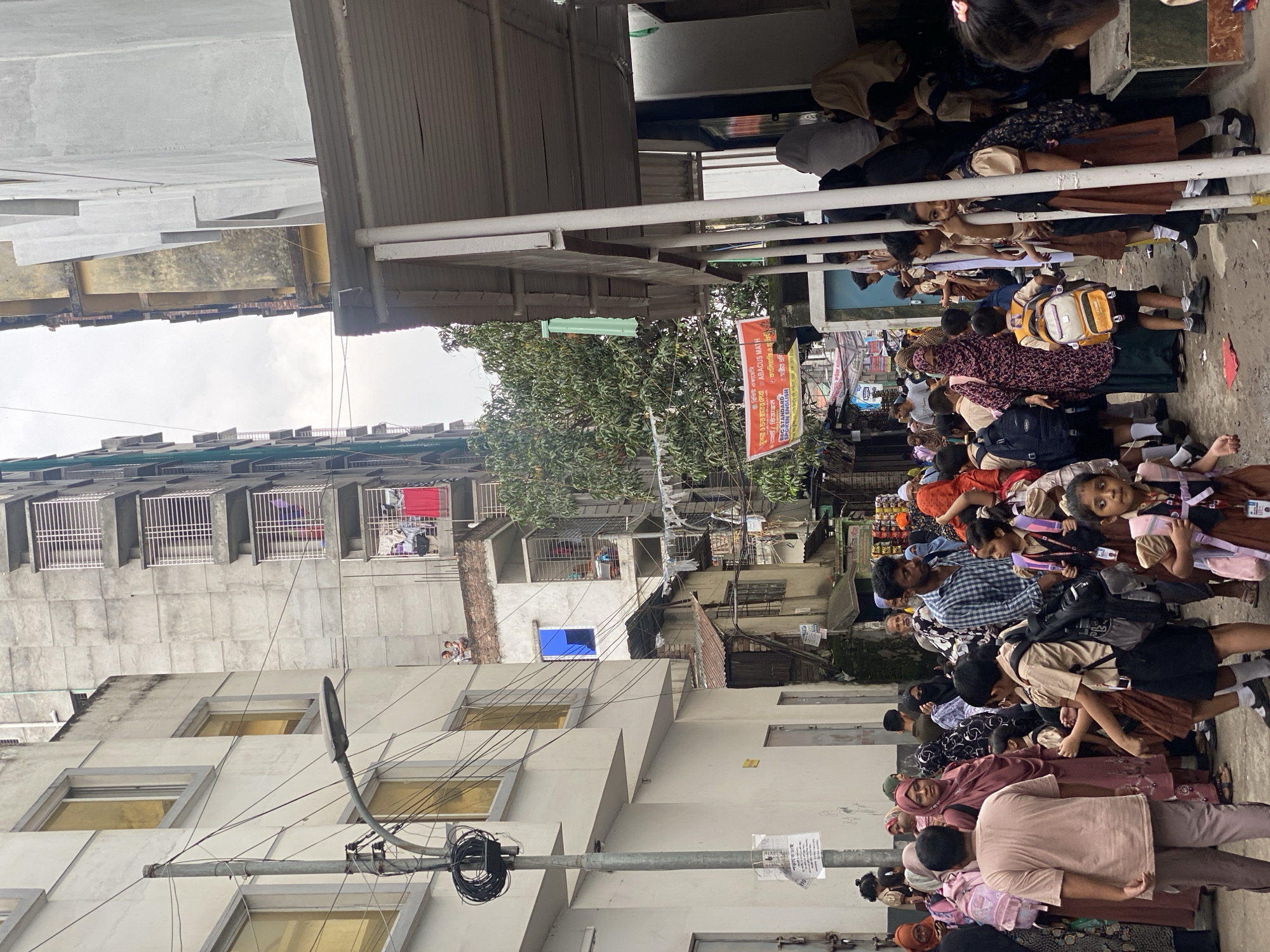 In an urban alleyway in Unknown, Unknown, a crowd of people, predominantly children in school uniforms, are gathered. The narrow space is flanked by multi-story buildings under construction or in disrepair, with exposed brick and concrete visible. Laundry hangs from some of the upper floors. A street lamp with coiled wires is a prominent fixture on the left. A red banner advertising educational services is hung across the street. Some children are wearing backpacks, suggesting they are either going to or returning from school. The overall atmosphere appears busy and somewhat crowded.