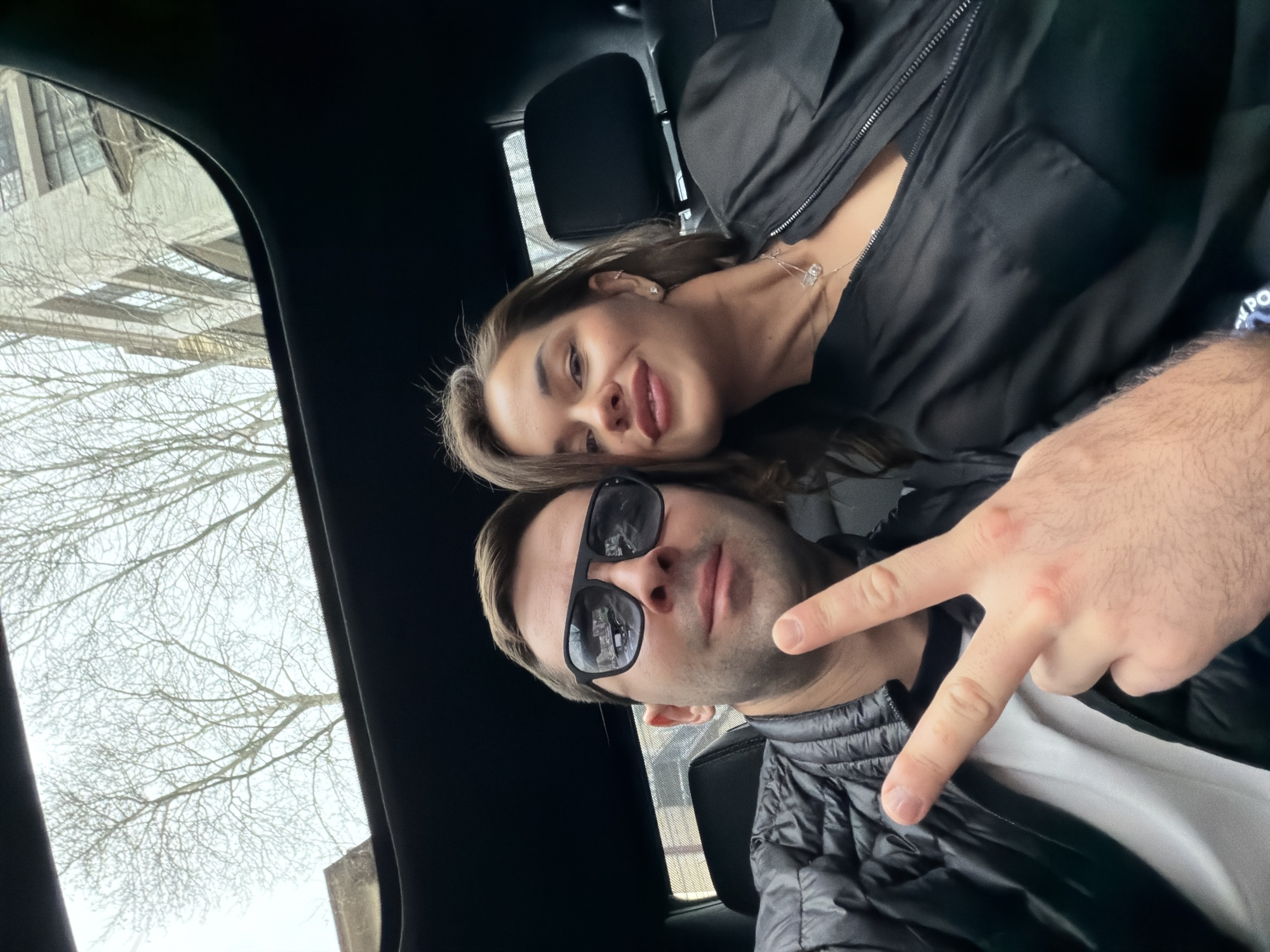 The image displays a selfie taken inside a vehicle, featuring two adult individuals. An adult male with short dark hair, stubble, and fair skin occupies the left side of the frame. He wears black rectangular sunglasses, a black quilted puffer jacket over a white t-shirt, and is seen from the upper torso. His right hand is raised, forming a V-sign with extended index and middle fingers. He looks directly at the camera with a slight smile. Adjacent to him on the right is an adult female with long brown hair and fair skin, wearing a black collared garment with a zipper and a silver-toned necklace featuring a clear pendant. She is observed from the shoulders up, smiling directly at the camera, with her head slightly angled towards the male. Both individuals are seated in dark-colored vehicle seats. The car's dark interior headliner and an open sunroof are visible above them. Through the sunroof, an overcast, light grey sky is apparent, interspersed with the bare, leafless branches of deciduous trees. A multi-story, light-colored urban building with windows and balconies is partially visible through the sunroof's upper right section. While the scene depicts an urban environment consistent with cities like Tbilisi, Georgia, no specific landmarks or unique identifiers are present in the image to pinpoint an exact location within the city.