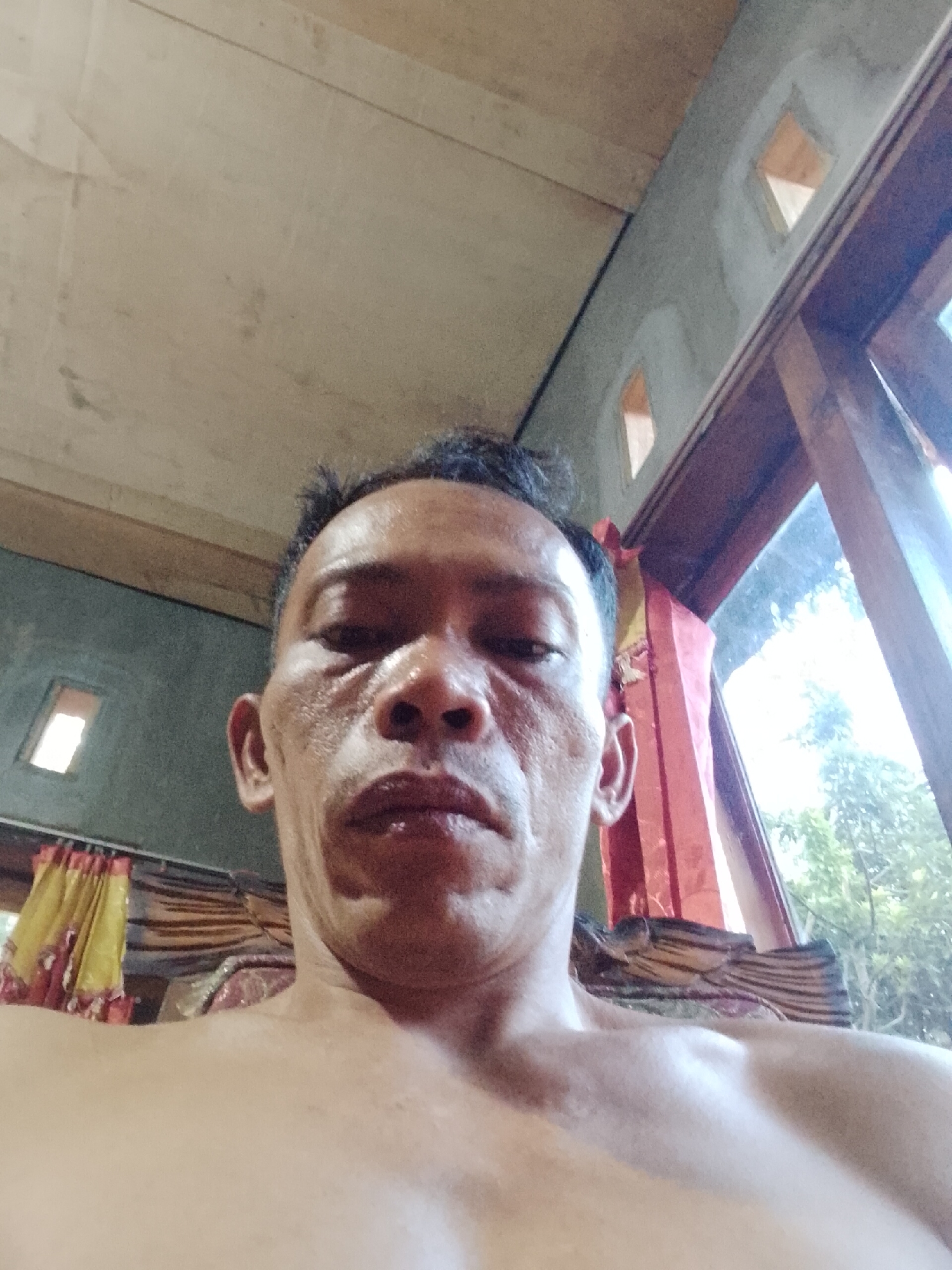 An over-the-shoulder, close-up, low-angle, selfie-style portrait depicts a shirtless man's face and upper chest. He has short, dark hair, prominent pores, and sun-damaged skin. His gaze is directed forward, with his mouth slightly open, revealing his lower teeth. The background features a ceiling made of wooden planks, a wall with small, diamond-shaped openings, and a window with wooden framing, partially obscured by red and yellow patterned curtains. Outside the window, trees and foliage are visible. The scene appears to be indoors, likely in a residential setting in Madukoro, Indonesia, given the architectural details and vegetation.