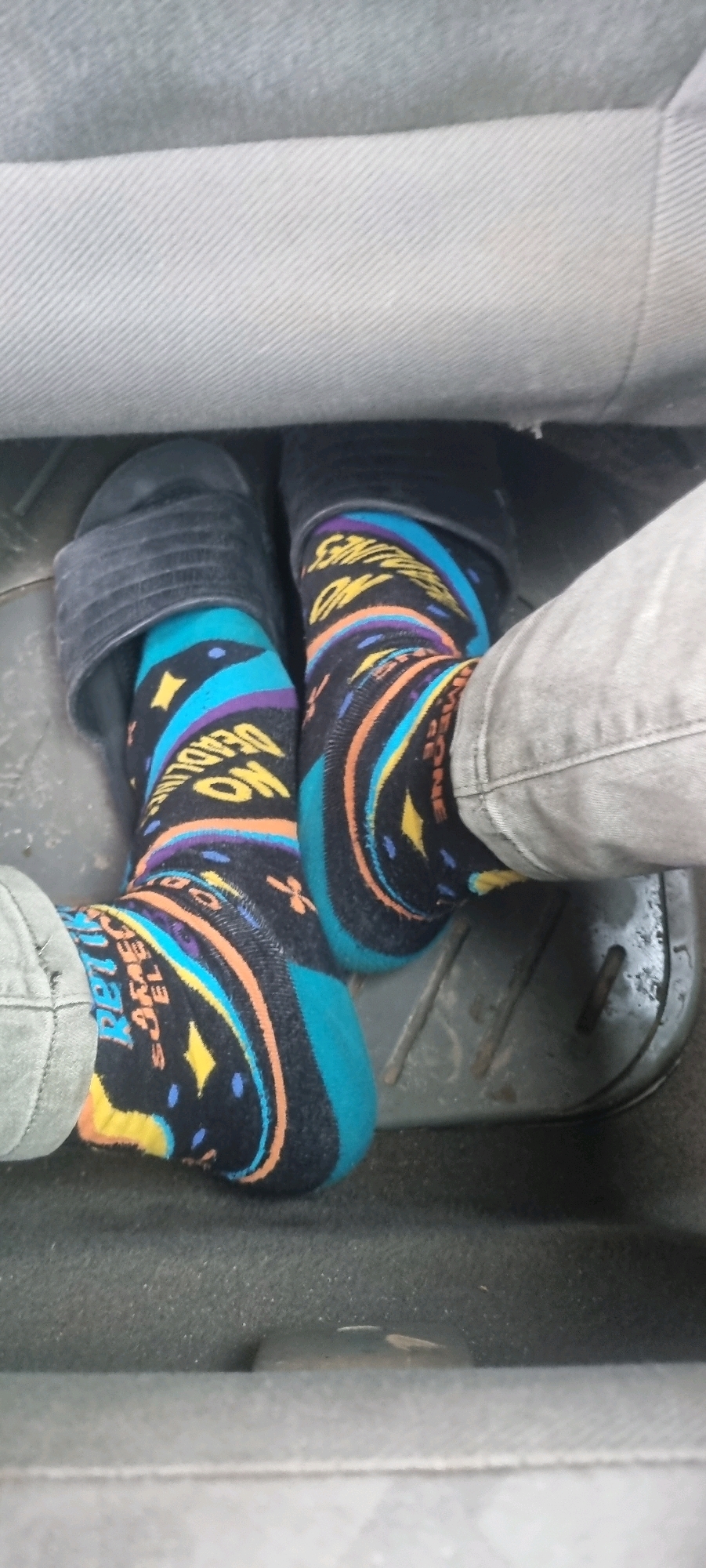 The image shows a close-up view of a person's feet wearing brightly colored socks with a geometric pattern and text. The person is wearing black sandals on the left foot and what appears to be a denim pant leg on the right leg, with the sock visible above the pant. The feet are positioned on a textured, dark gray floor mat, likely inside a vehicle, as suggested by the presence of a gray fabric surface in the background which could be a car seat. One sock has the text "NO DEADLINES" printed on it in yellow. The other sock has text that is partially obscured, but appears to include "AWESOME". The overall scene is indoors and the lighting suggests it is daytime. This image does not contain information about the city of Iju, Nigeria.