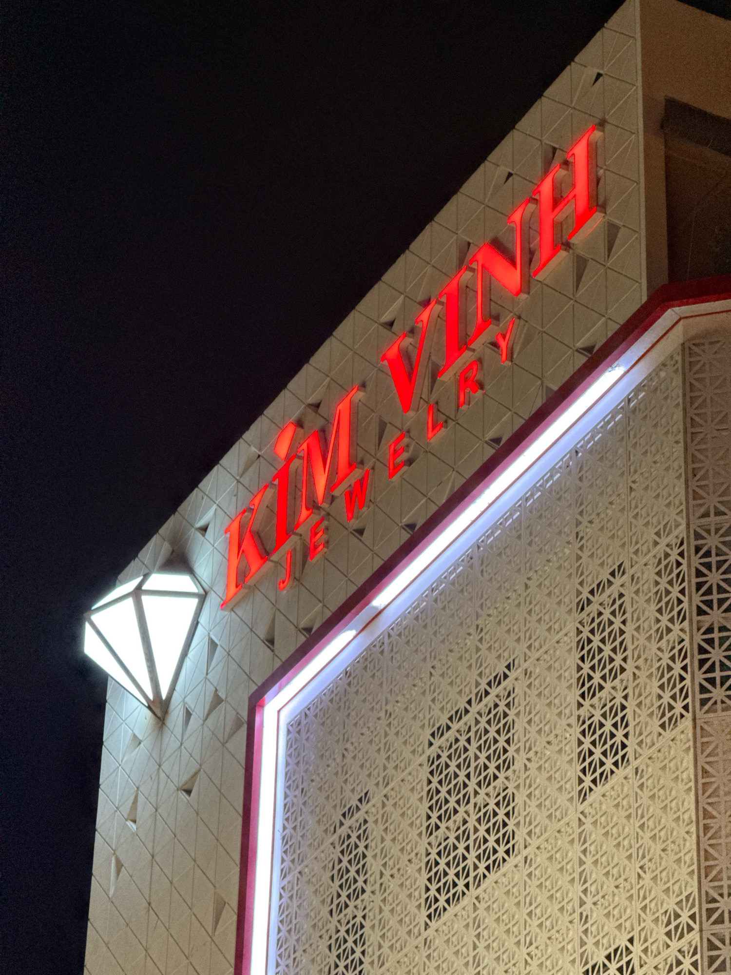The image shows the exterior of a building at night, featuring a prominent red neon sign that reads "KIM VINH JEWELRY". The building's facade is decorated with a textured pattern of small triangles. To the left of the sign, a stylized diamond-shaped light fixture is visible. The lower part of the building displays a decorative screen with intricate geometric patterns. The scene is lit by the neon sign and ambient lighting from within the building. The location is identified as Nha Trang, Vietnam.