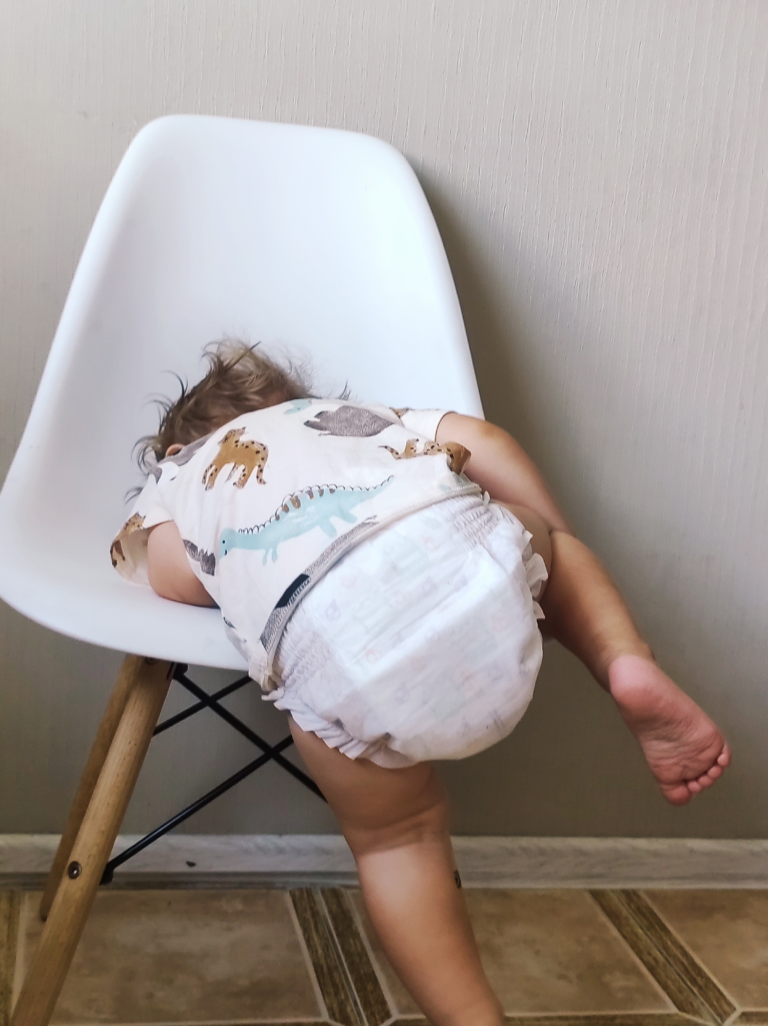 A child wearing a diaper and a shirt with a dinosaur print is sleeping slumped over a white chair. The child's head is resting on their arms, and their legs are hanging over the side of the chair. The chair has wooden legs and a black metal frame. The background is a plain, textured wall and a tiled floor. The scene appears to be indoors, possibly in a home, in the city of Chaikovsky, Russia.