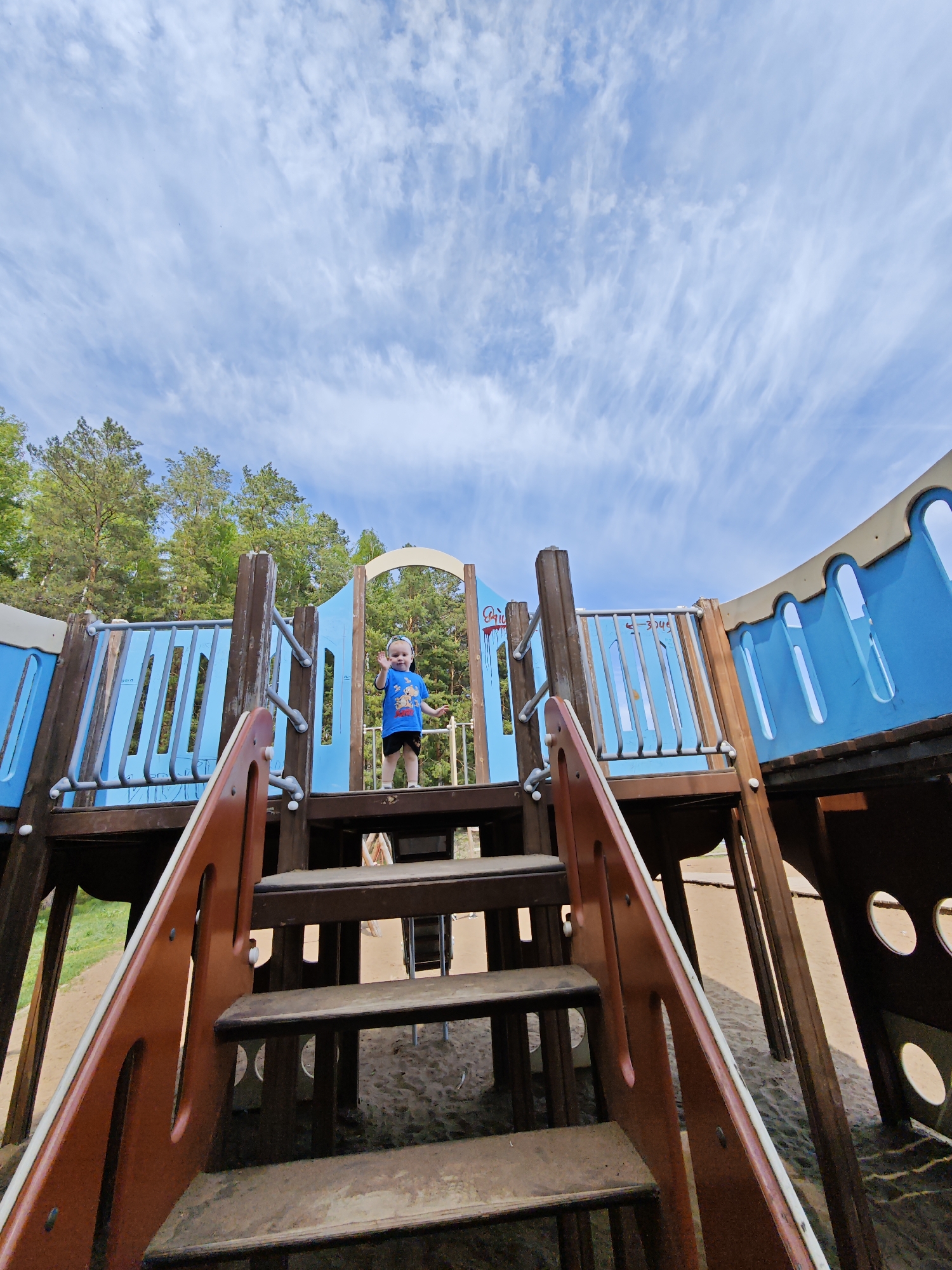 A low-angle, wide-lens shot of a playground in Chaikovsky, Russia, features a toddler standing atop a section of the structure, waving to the camera. The playground is primarily constructed of wood, painted in shades of blue and brown, and includes a set of stairs in the foreground leading up to the toddler's position. The sky is visible in the background, filled with wispy white clouds. Trees are partially visible behind the playground structure. The area beneath the playground appears to be covered in sand.