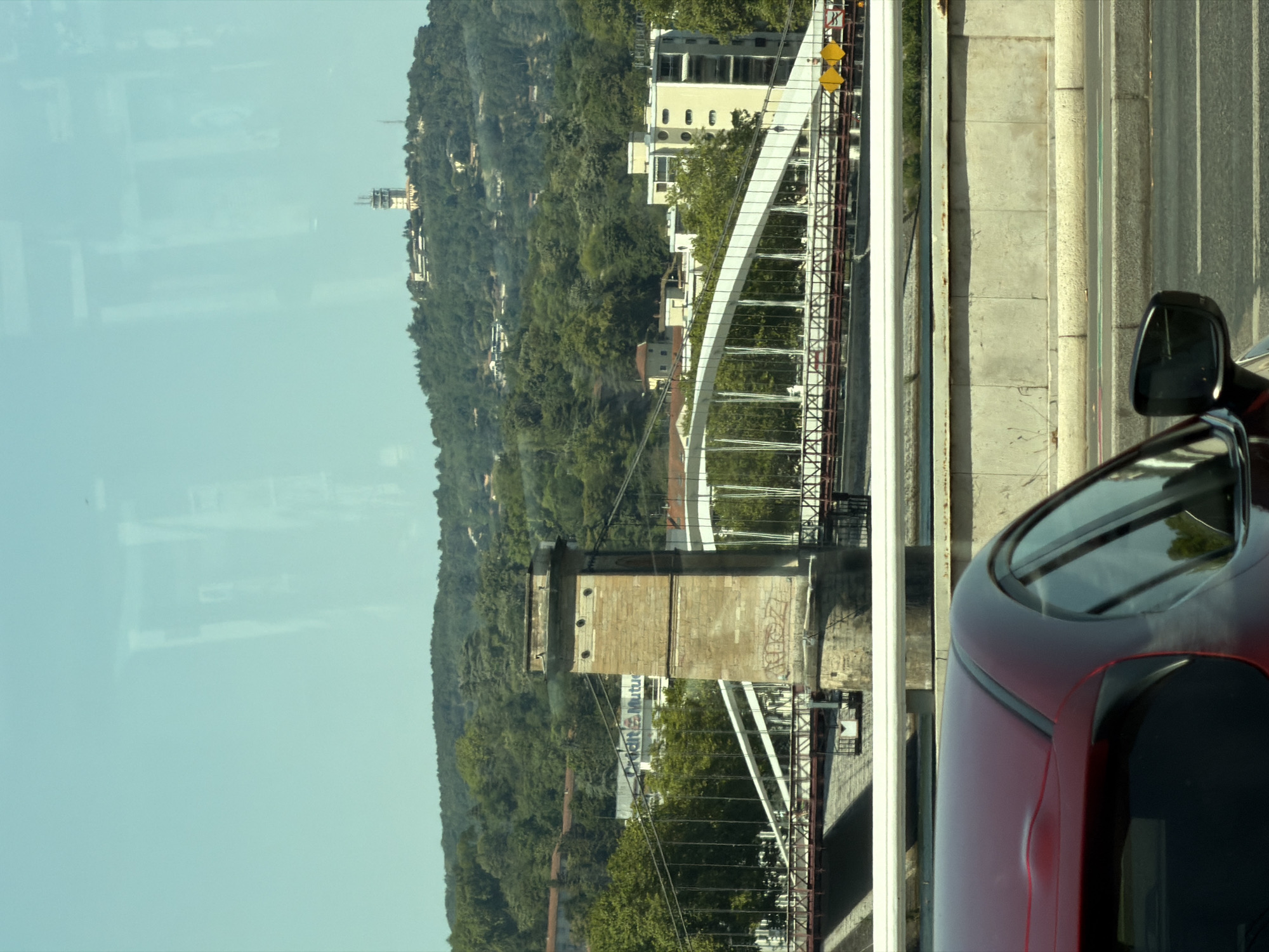 The image captures a view of the Pont de la Mulatière in Lyon, France, seen from a moving vehicle. In the foreground, the rear portion of a red car is visible, with its roof and side mirror. Beyond the car, a concrete barrier and a section of road are present. The middle ground features the Pont de la Mulatière, a suspension bridge with white arches supported by stone towers. A sign for "Crédit Mutuel" is visible behind the bridge. In the background, a lush green hillside is covered in trees, with buildings scattered across it. A tall structure, possibly a radio tower or observation deck, stands atop the hill. The sky is a clear blue. Reflections of what appear to be buildings or structures are present on the glass through which the photograph was taken.
