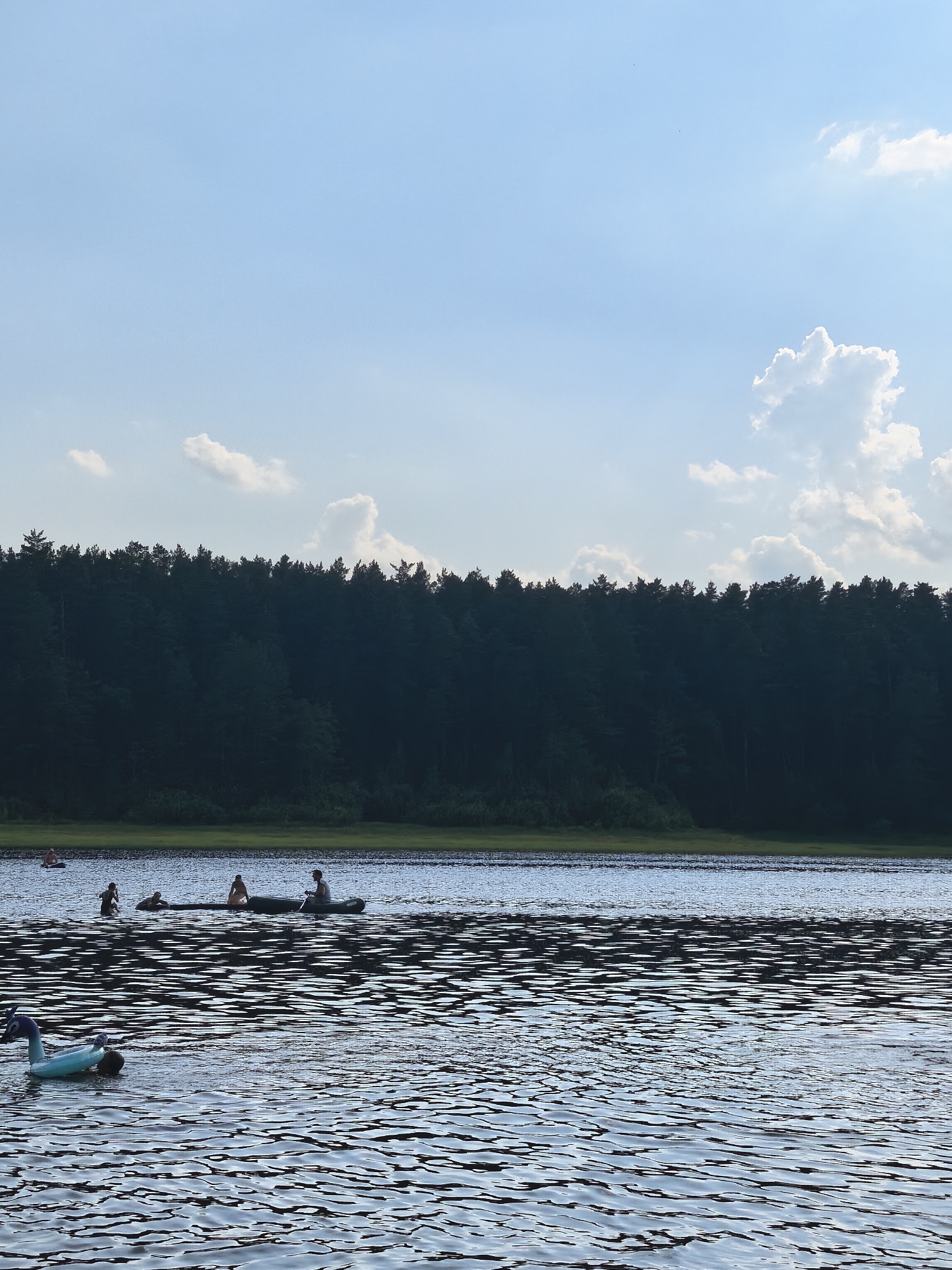 This media file captures a serene summer scene on a lake in Sysert, Russia. The image is dominated by the calm, rippling water, reflecting the clear blue sky dotted with fluffy white clouds. In the distance, a dense line of dark green trees forms a natural backdrop to the tranquil setting. The time of day appears to be late afternoon or early evening, as indicated by the soft lighting and the slight haze in the air.
Several individuals are enjoying recreational activities on the water. In the foreground, an inflatable blue toy, possibly a pool float, is partially submerged. Further out on the lake, a group of people are engaged in various water activities. One group is in a canoe, with at least two individuals paddling, while another person sits in the boat. Nearby, other individuals are swimming or wading in the water. The atmosphere seems relaxed and pleasant, suggesting a leisurely day spent outdoors. The overall impression is one of peaceful enjoyment of nature's beauty. There is no visible text in the image.